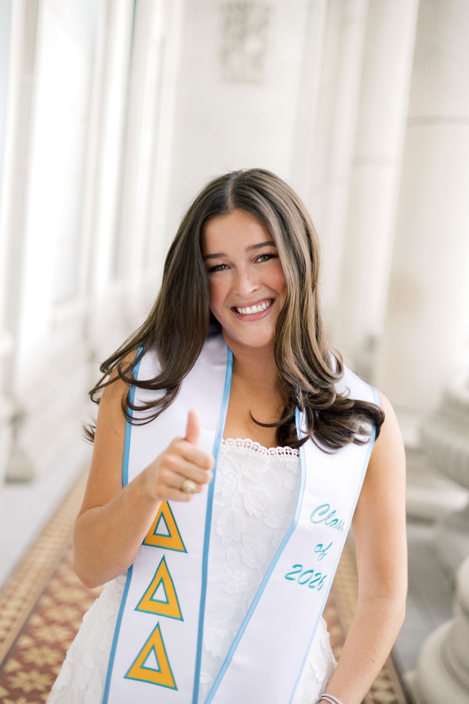 Smiling Tri Delta Class of 2024 graduate in white lace dress gives thumbs up wearing sorority stole.