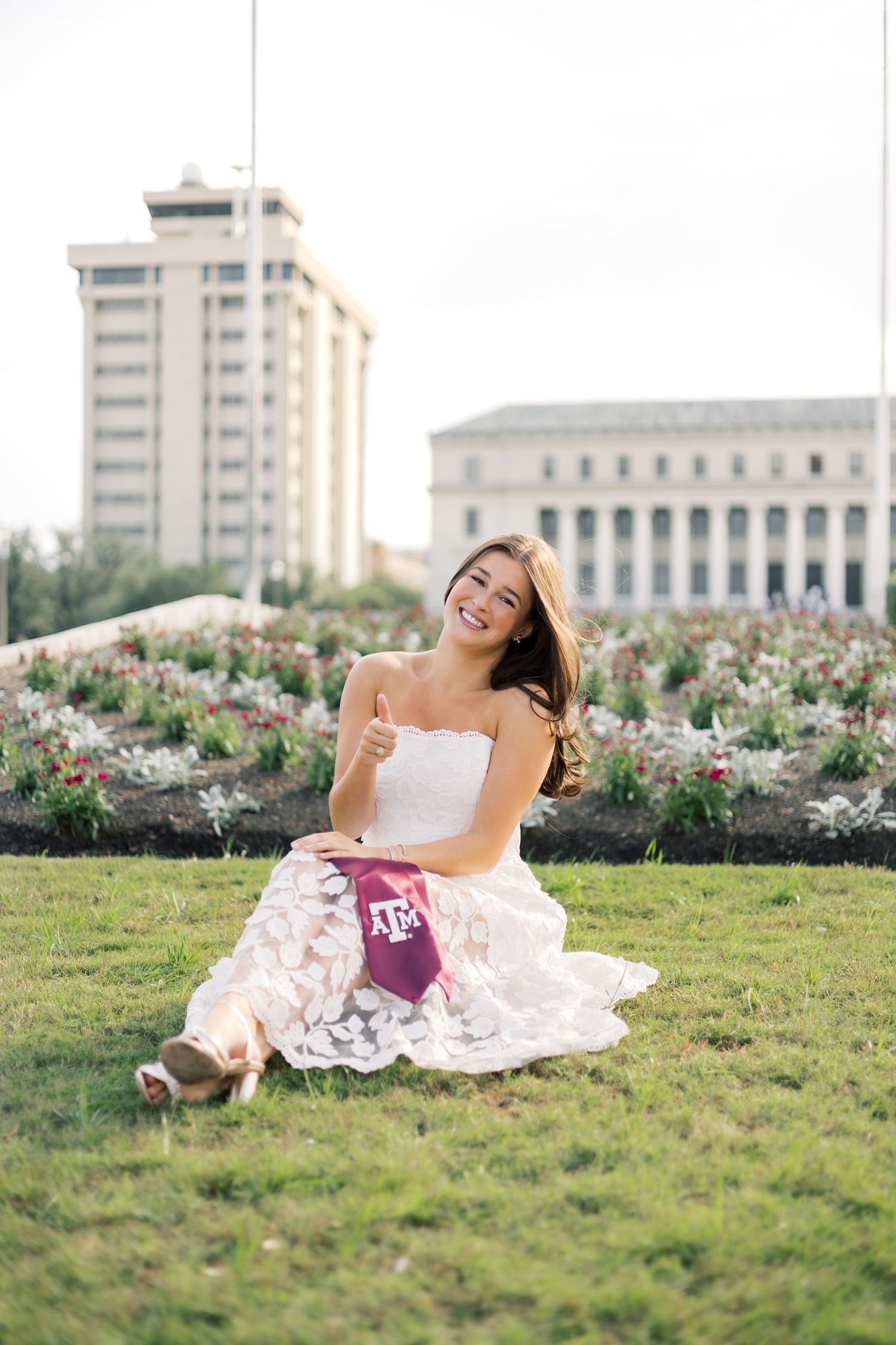 Happy college graduate in white lace dress sitting on grass giving thumbs up at Texas A&M University.