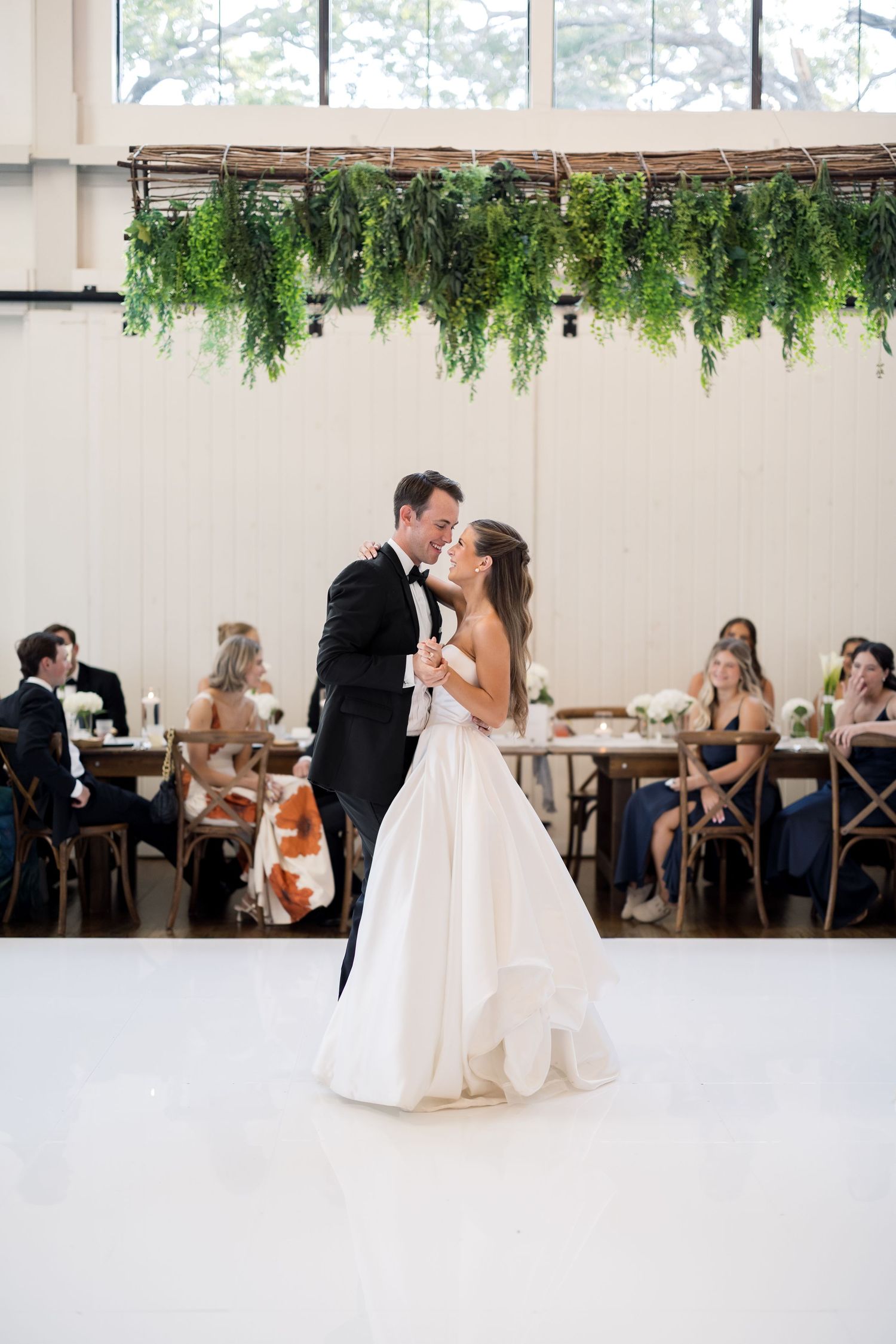 Bride and groom share first dance on white floor beneath lush greenery installation at elegant wedding reception.
