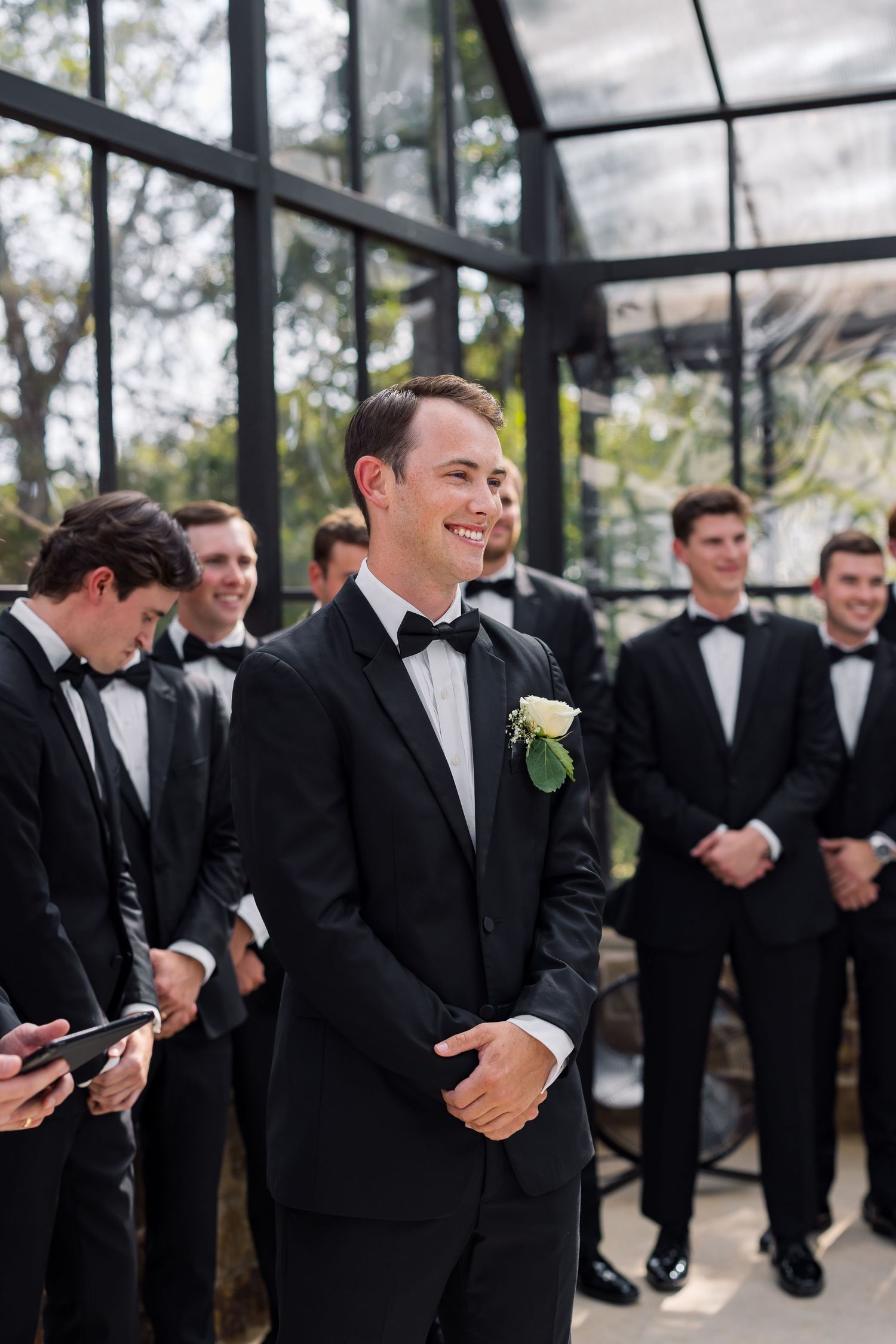 Smiling groom in black tuxedo with white boutonniere stands at altar with groomsmen lined up behind him at wedding ceremony.