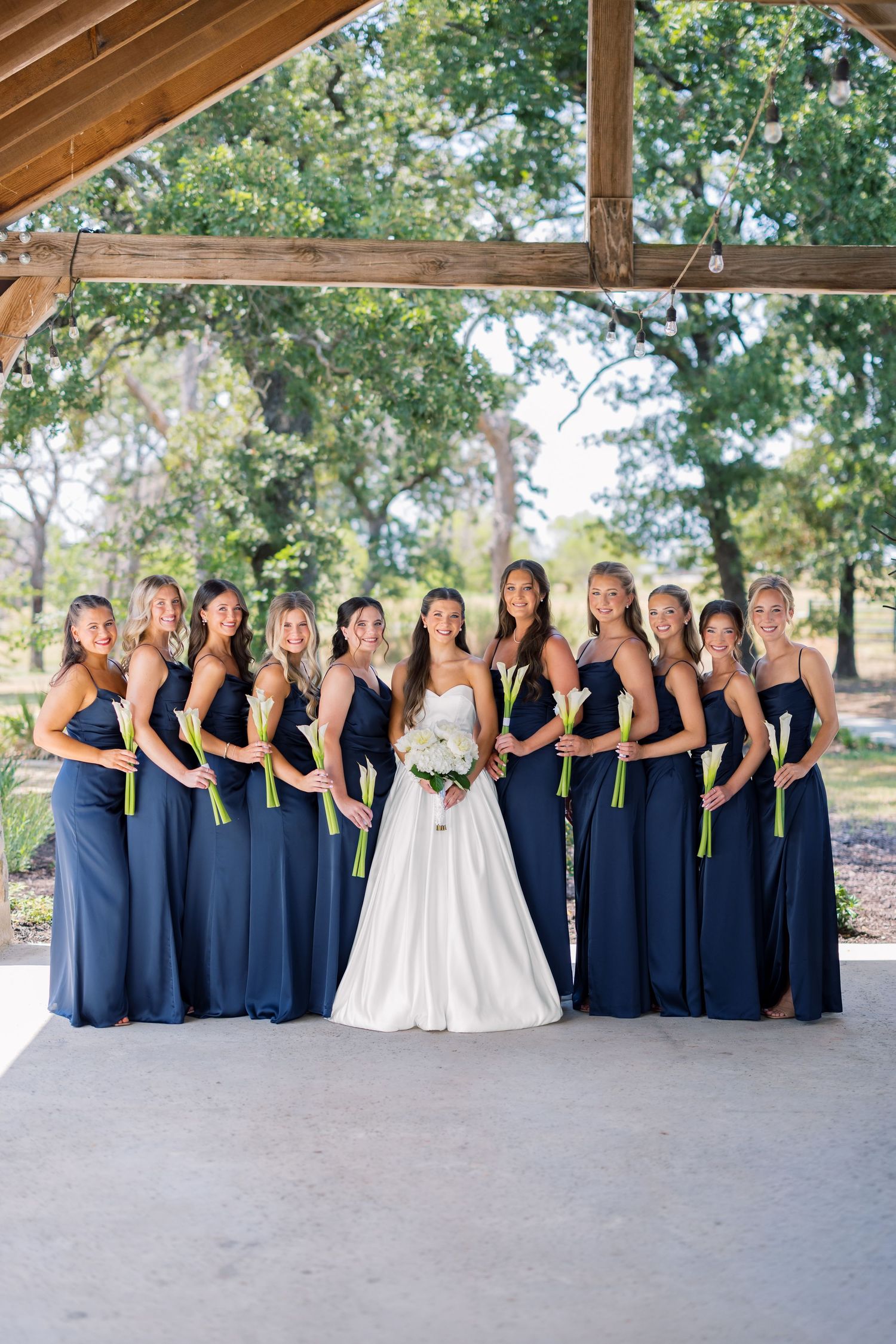 Bride in white gown poses with ten bridesmaids in navy blue dresses holding green-stemmed bouquets outdoors.