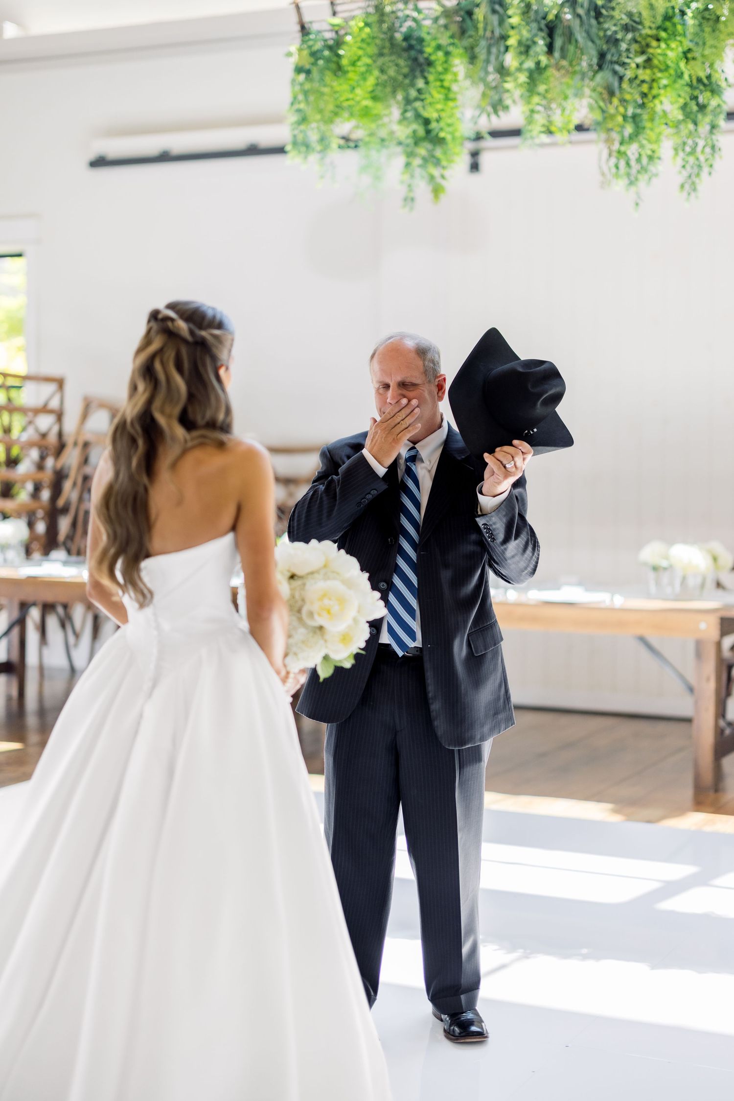 Emotional father holds hat to chest as bride in white gown holds bouquet during wedding ceremony indoors.