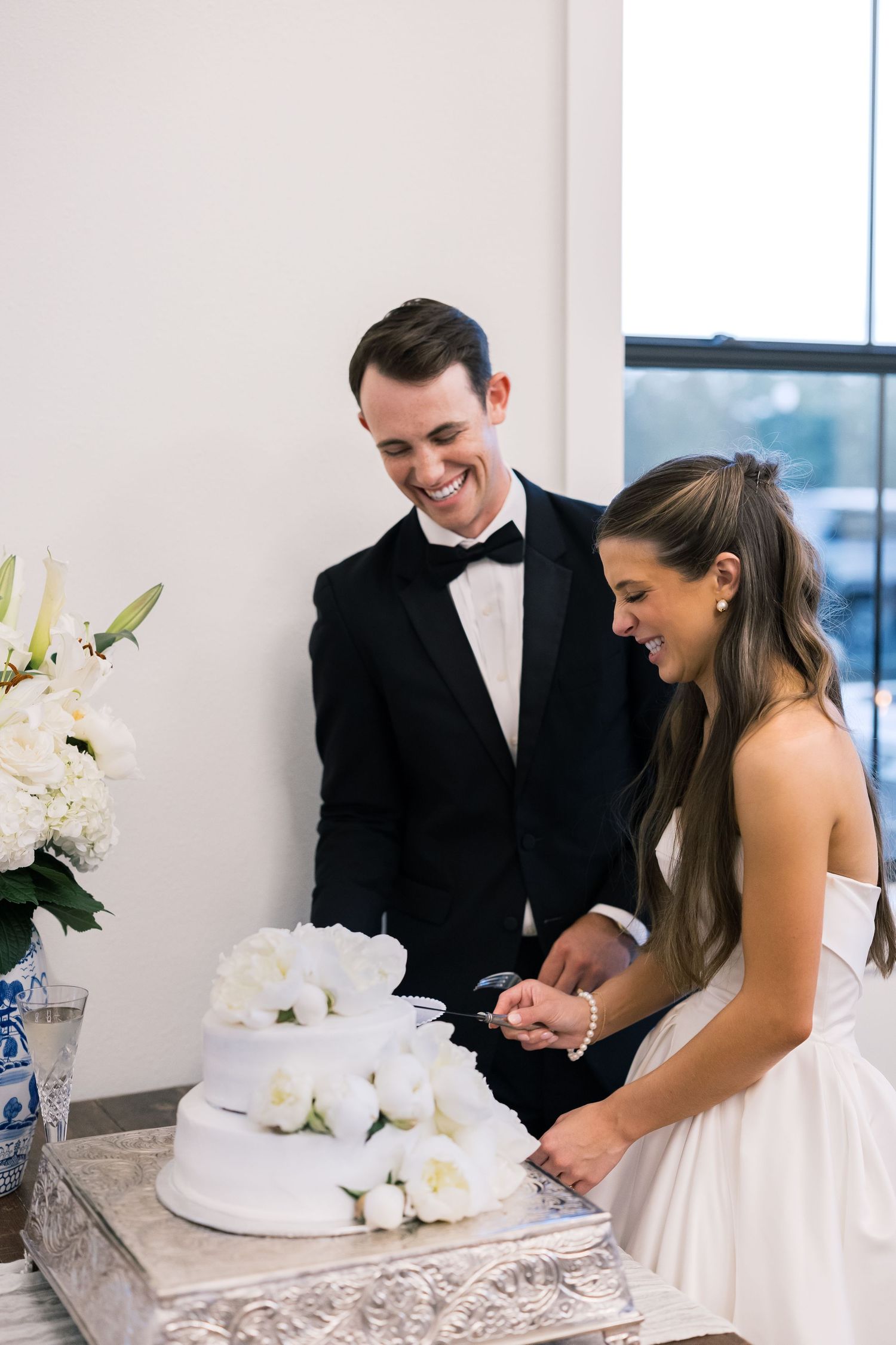 Smiling bride and groom cutting white floral wedding cake at elegant reception table with white lily arrangement.