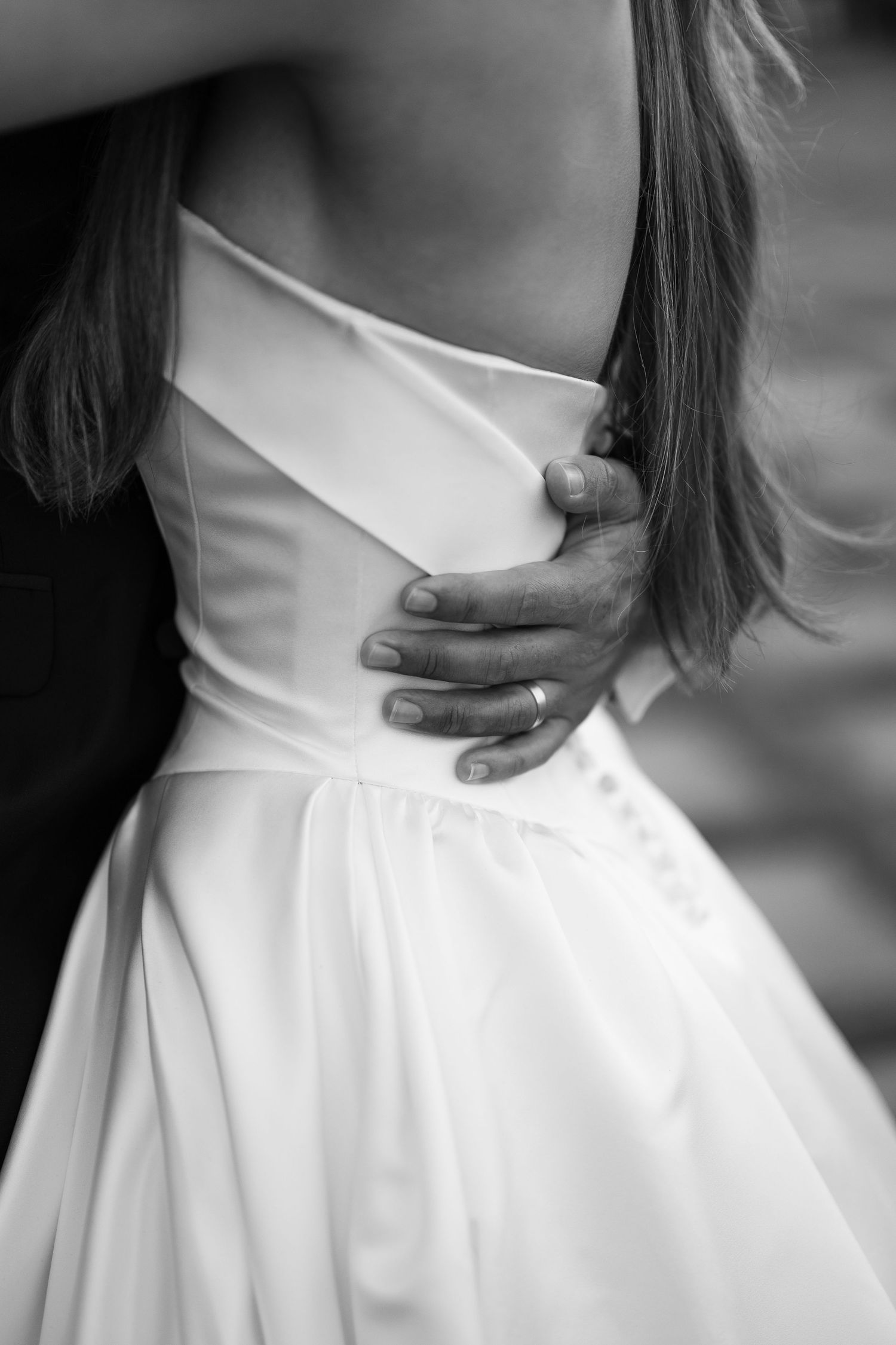 Black and white photo of a groom's hand with wedding ring embracing a bride in a white satin wedding dress.