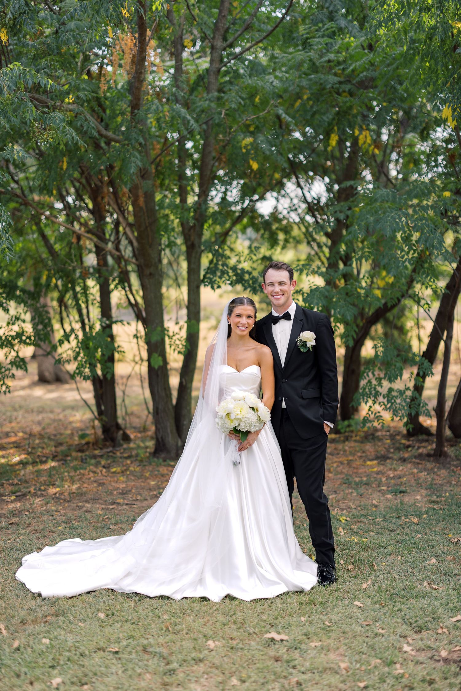 Bride in white strapless gown and groom in black tuxedo pose together outdoors among green trees on their wedding day.
