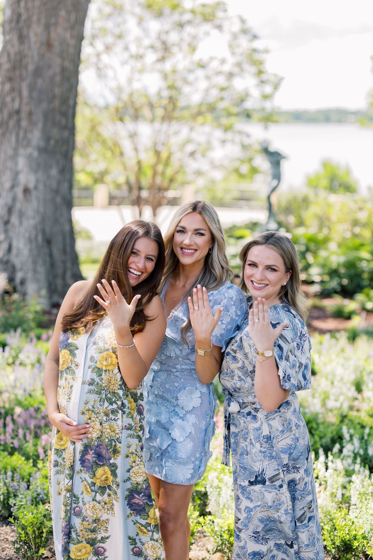 Three women in floral dresses posing in a garden, showing off their rings while smiling outdoors near a lake.