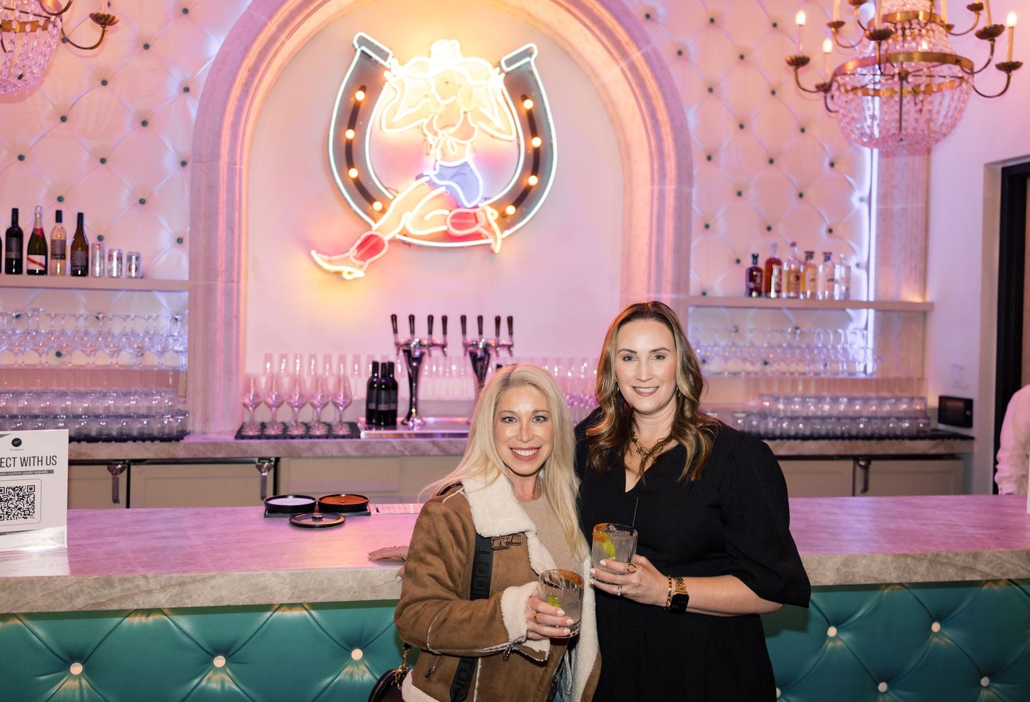 Two women smiling at a stylish bar with teal booths and a neon horseshoe sign backdrop.