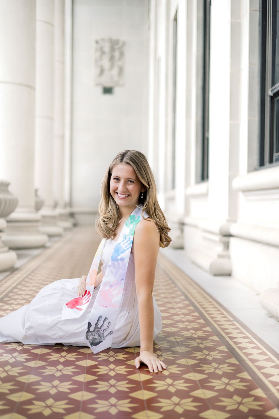 Smiling young woman in colorful handprint dress sitting on ornate tiled floor between white columns.