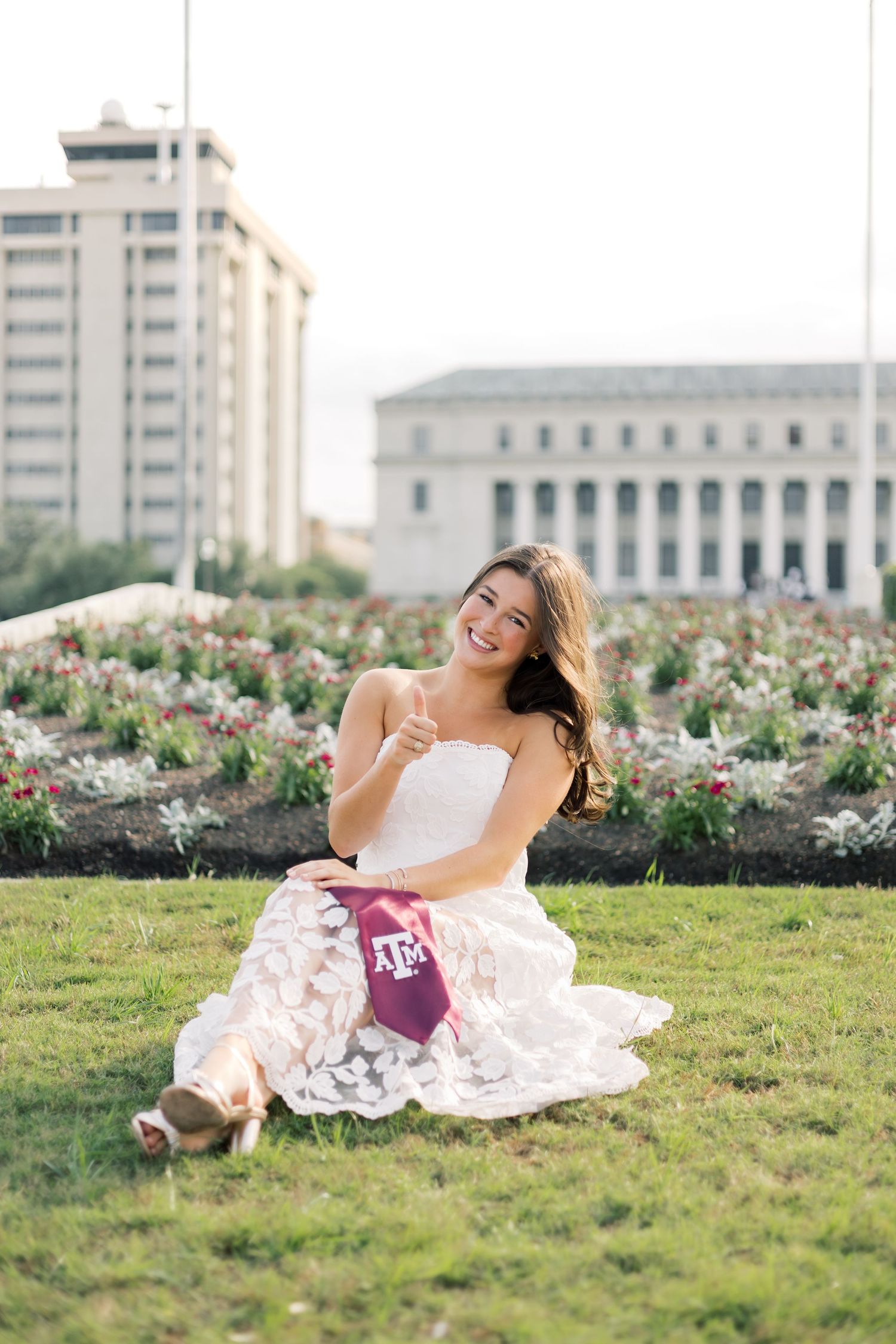 Happy woman in white strapless lace dress holding Texas A&M maroon stole, sitting on grass with campus in background.