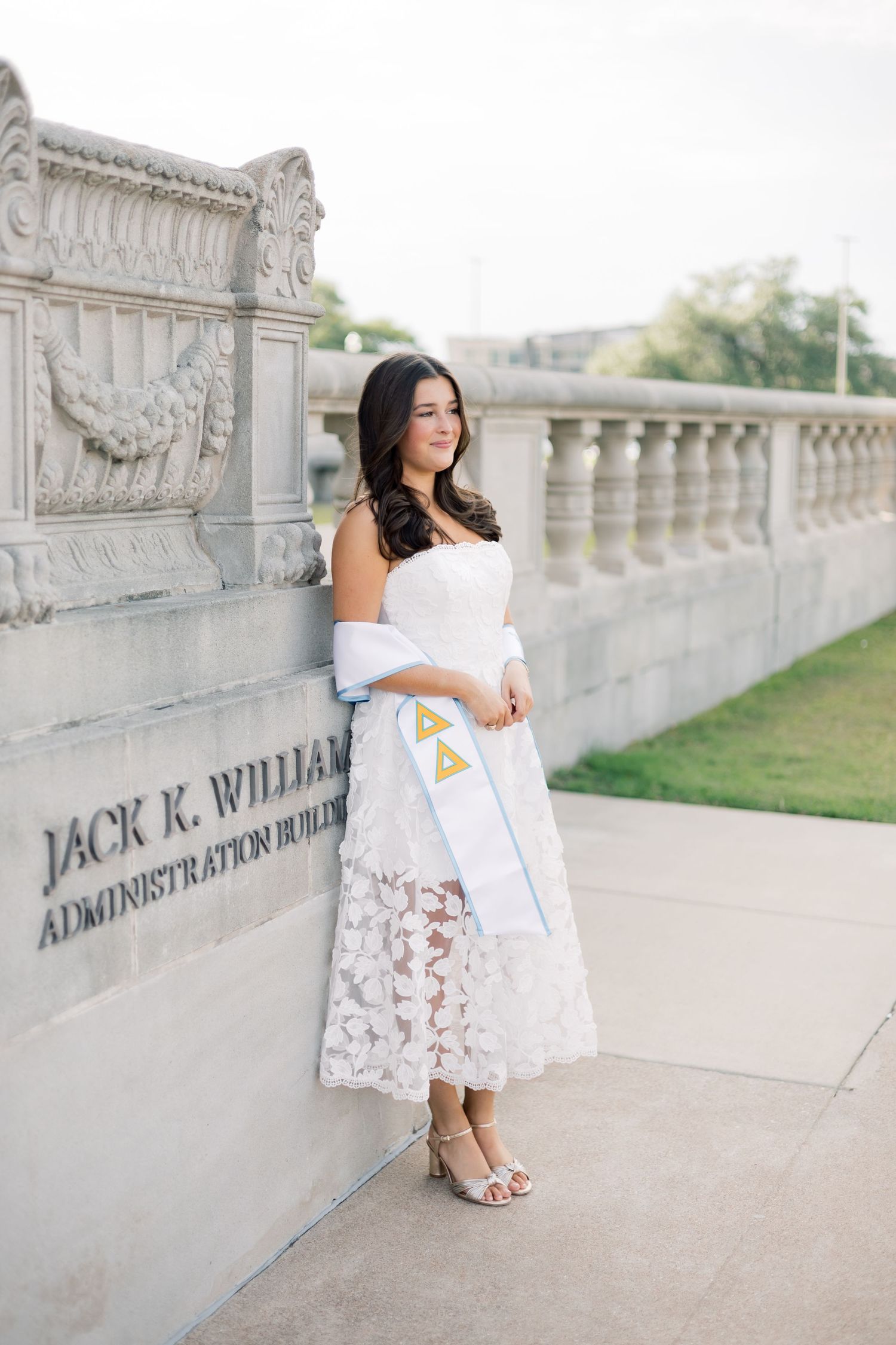 Graduate in white lace dress leaning against Jack K. Williams Administration Building with tri-delta sash.
