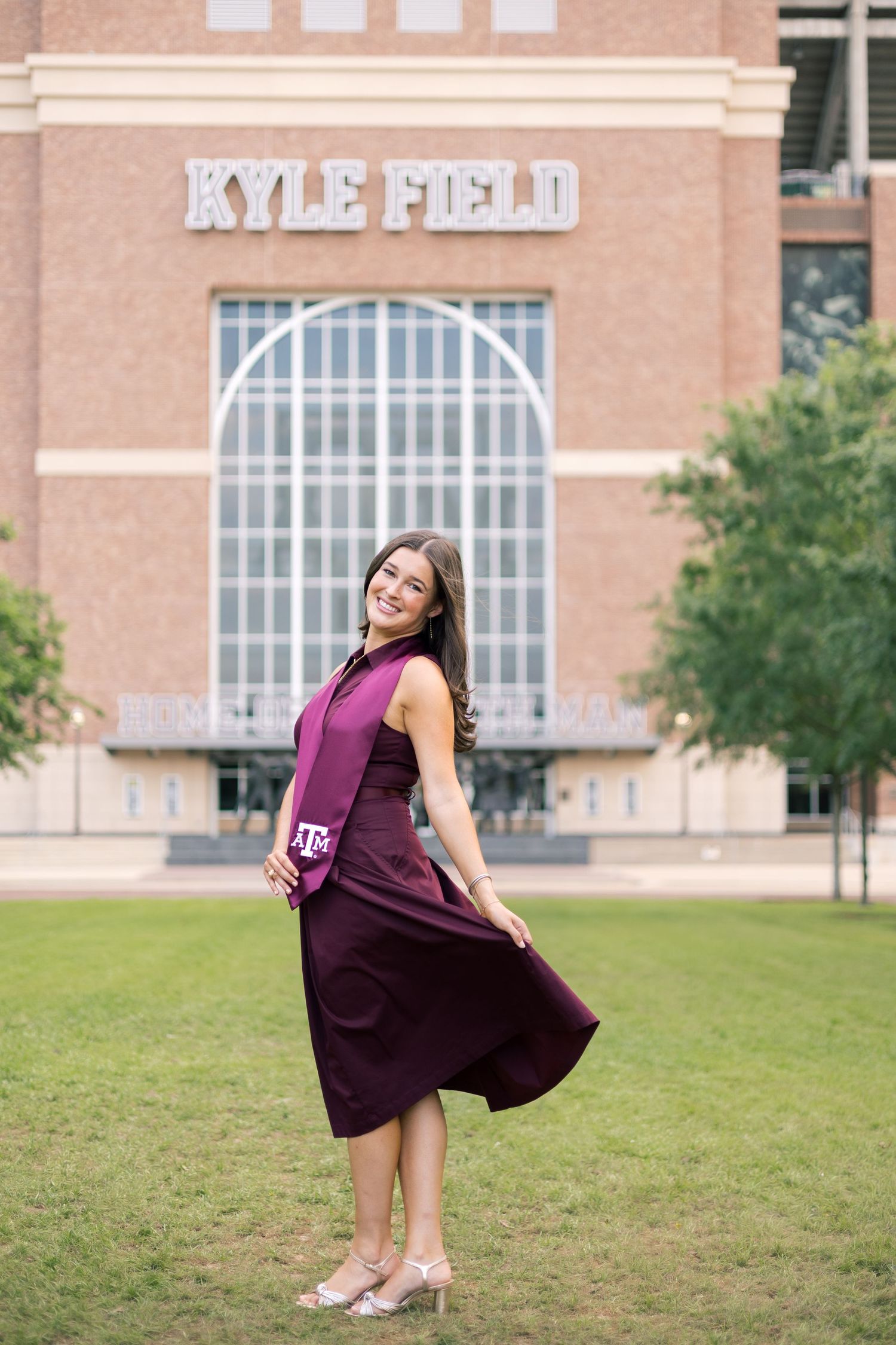 Texas A&M graduate twirls in maroon dress on green lawn in front of Kyle Field stadium.