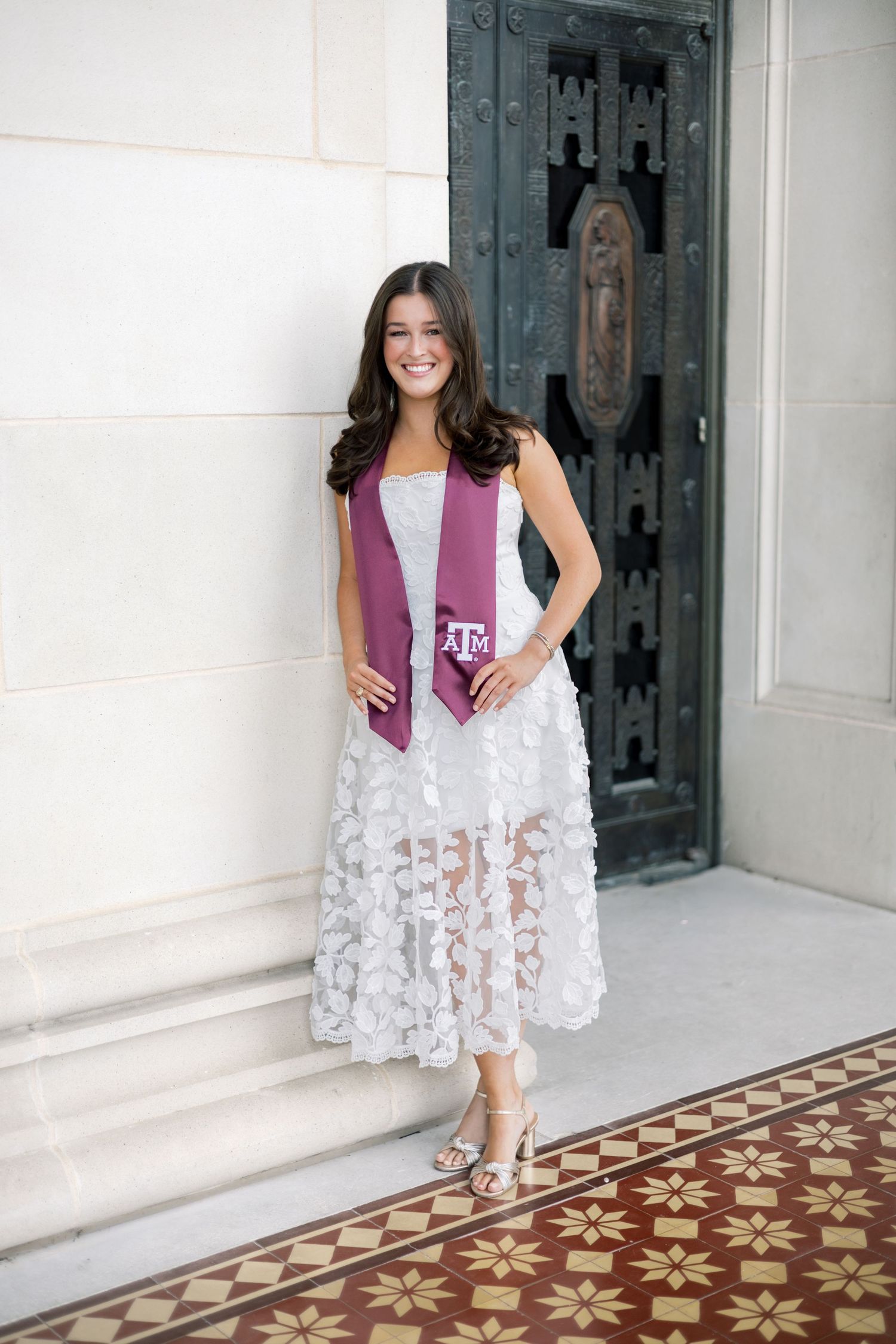 Texas A&M graduate in white floral lace dress and maroon stole stands smiling by decorative building entrance.