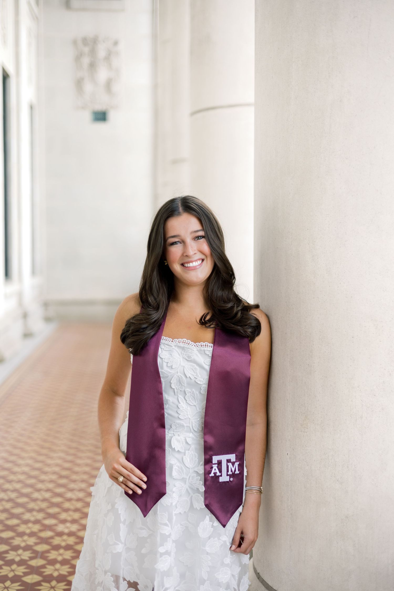 Smiling Texas A&M graduate in white lace dress with maroon ATM stole leans against white column.