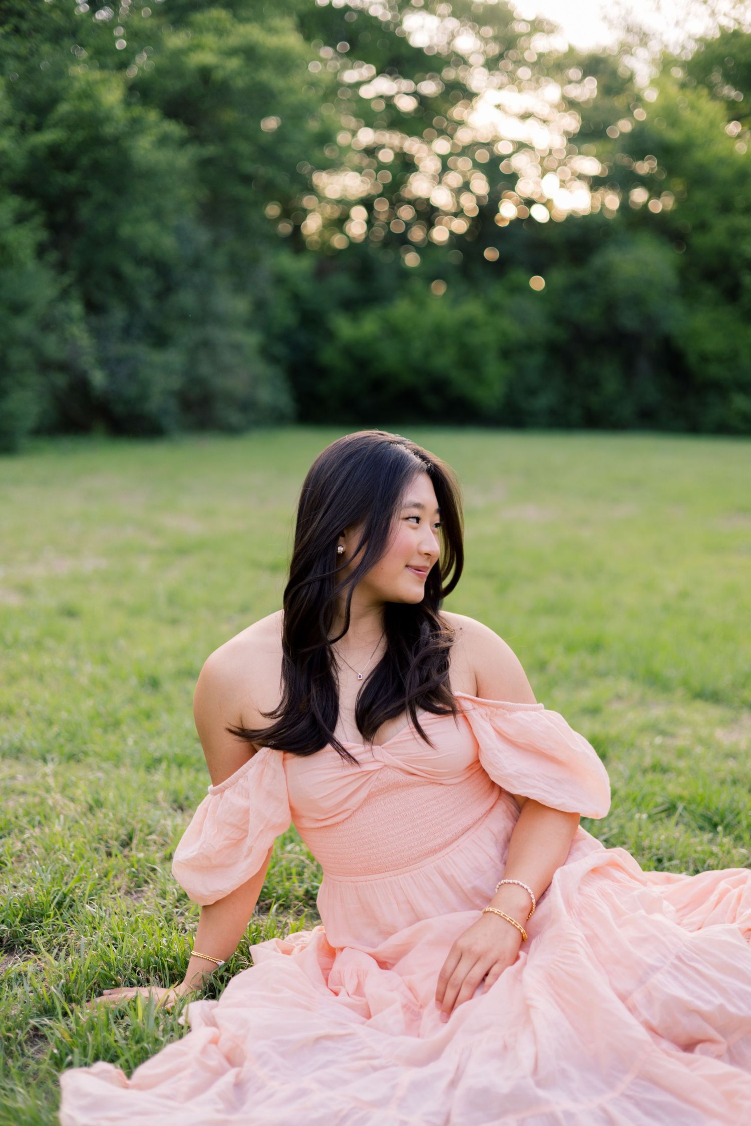 Young woman in pink off-shoulder dress sitting gracefully on green grass in a sunlit park.