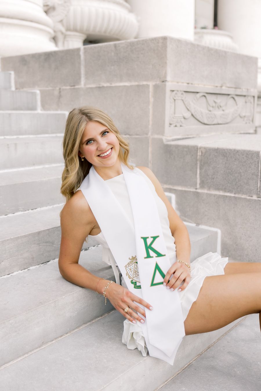 Smiling blonde woman sitting on stone steps wearing a white Kappa Delta sorority graduation stole.