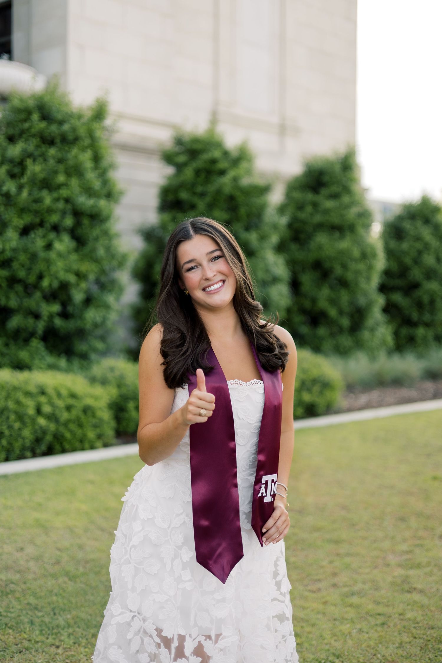 Smiling Texas A&M graduate in white lace dress giving thumbs up with maroon Aggie graduation stole.