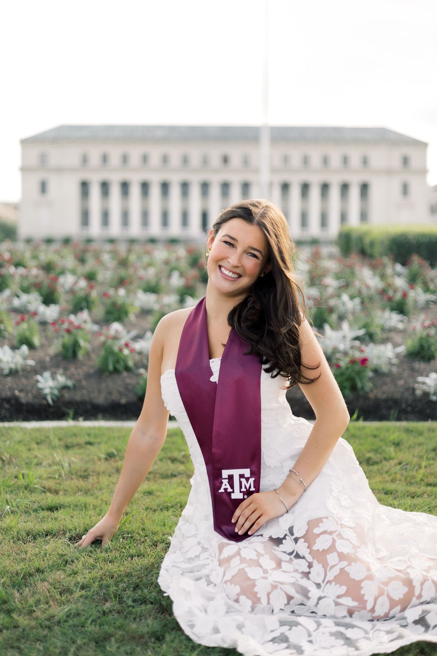 Joyful woman in white lace dress wearing Texas A&M maroon graduation stole seated on grass with campus building behind.
