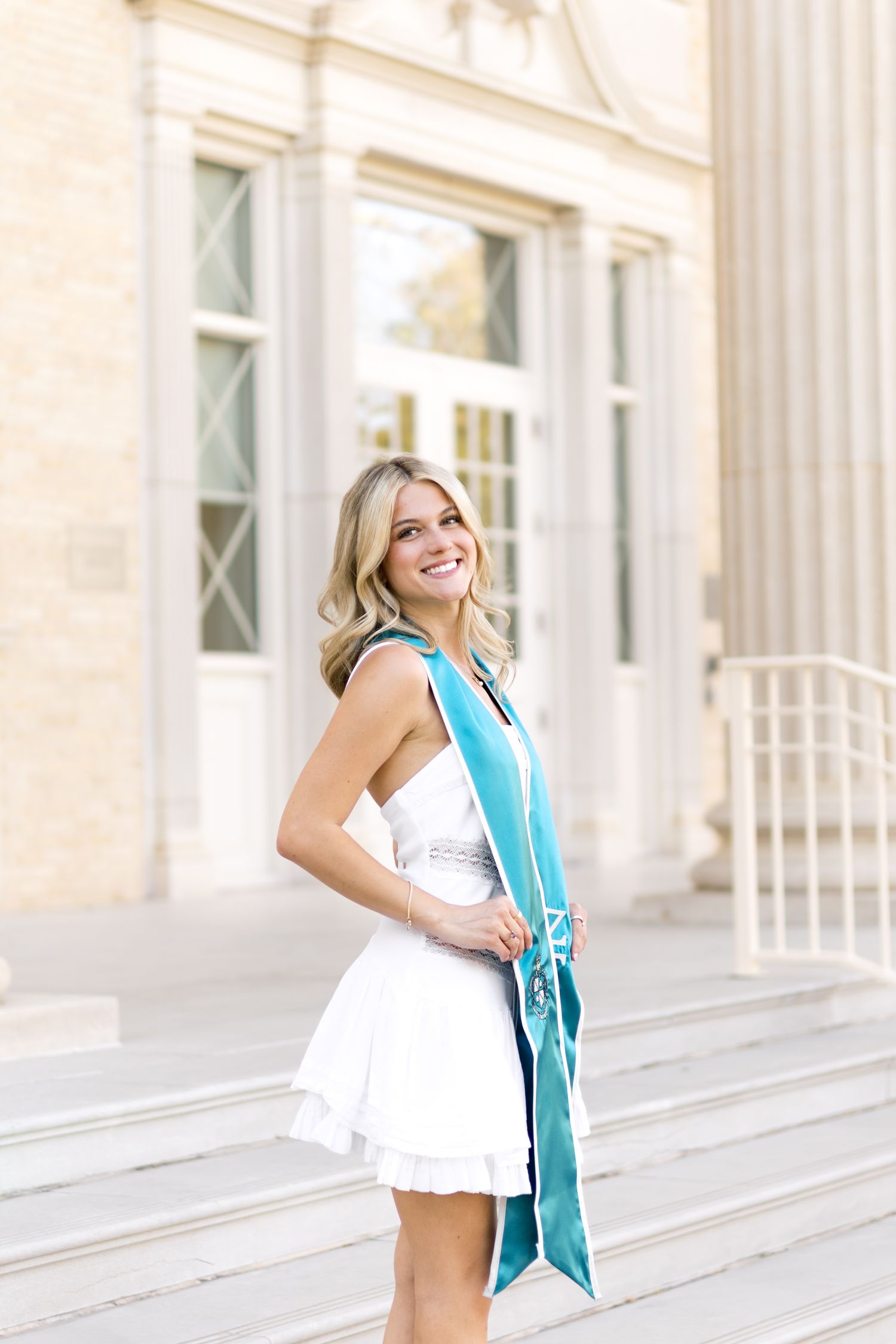 Smiling blonde woman in white dress wearing teal graduation sash on steps of TCU building.
