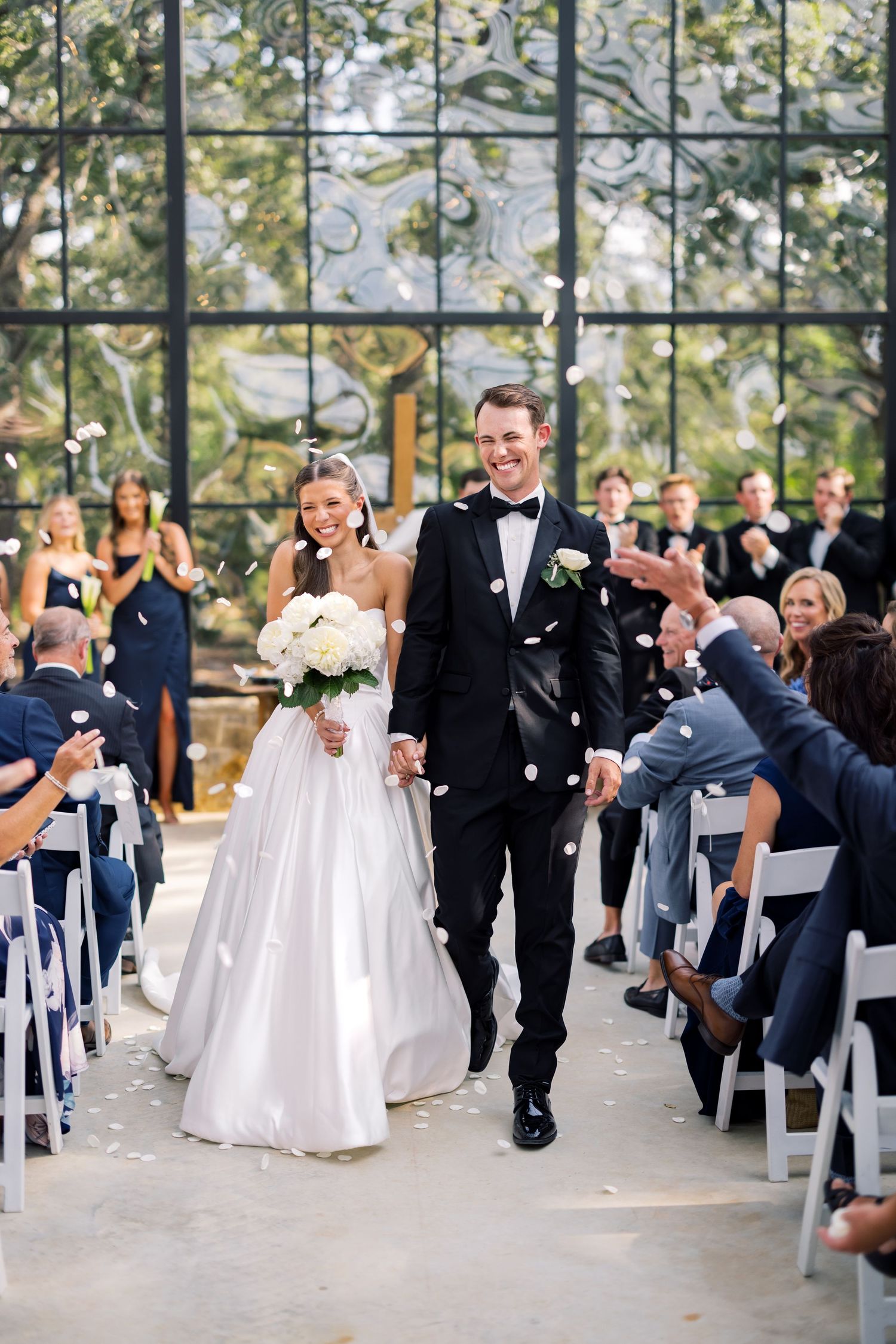 Bride and groom walk down the aisle as guests toss flower petals in an elegant glass chapel.