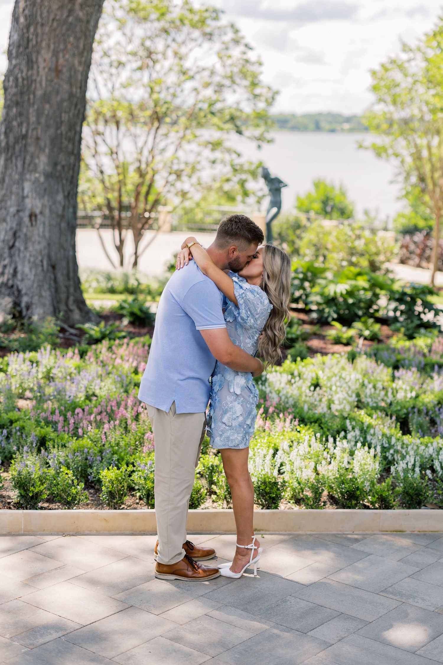 Couple sharing a romantic kiss in a lush garden with colorful flowers, a statue, and a serene lake in the background.