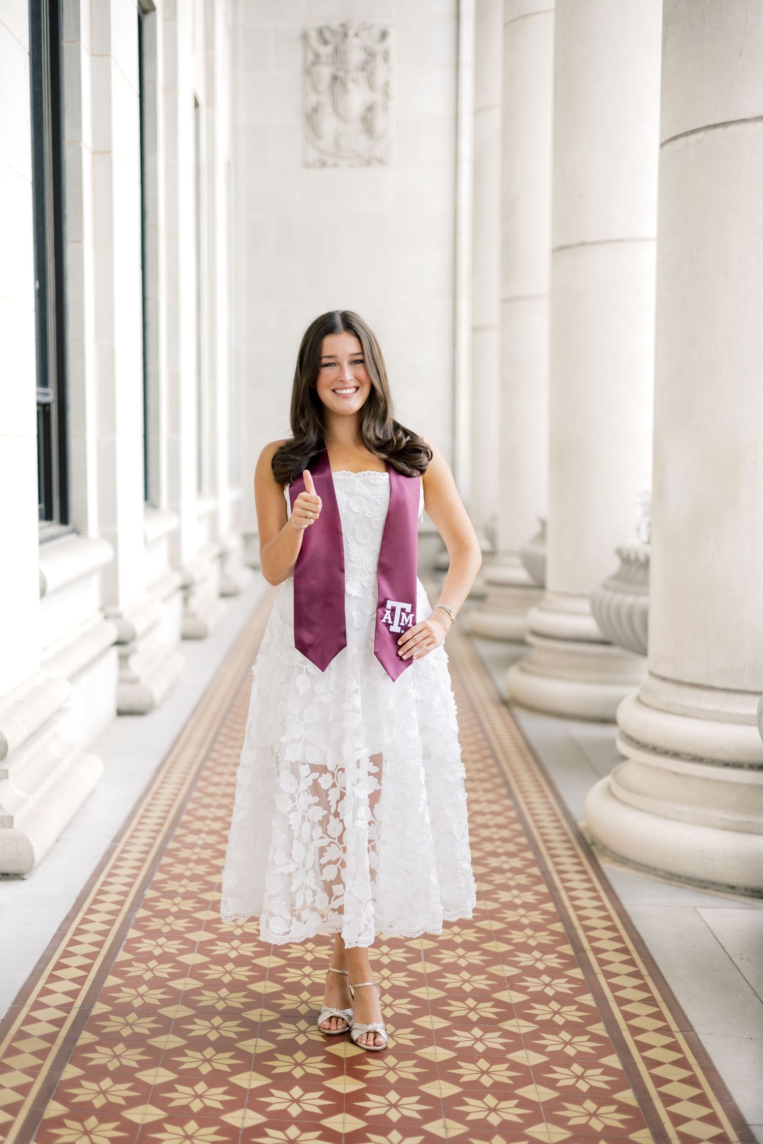 Texas A&M graduate in white lace dress gives thumbs up wearing maroon stole in elegant columned hallway.