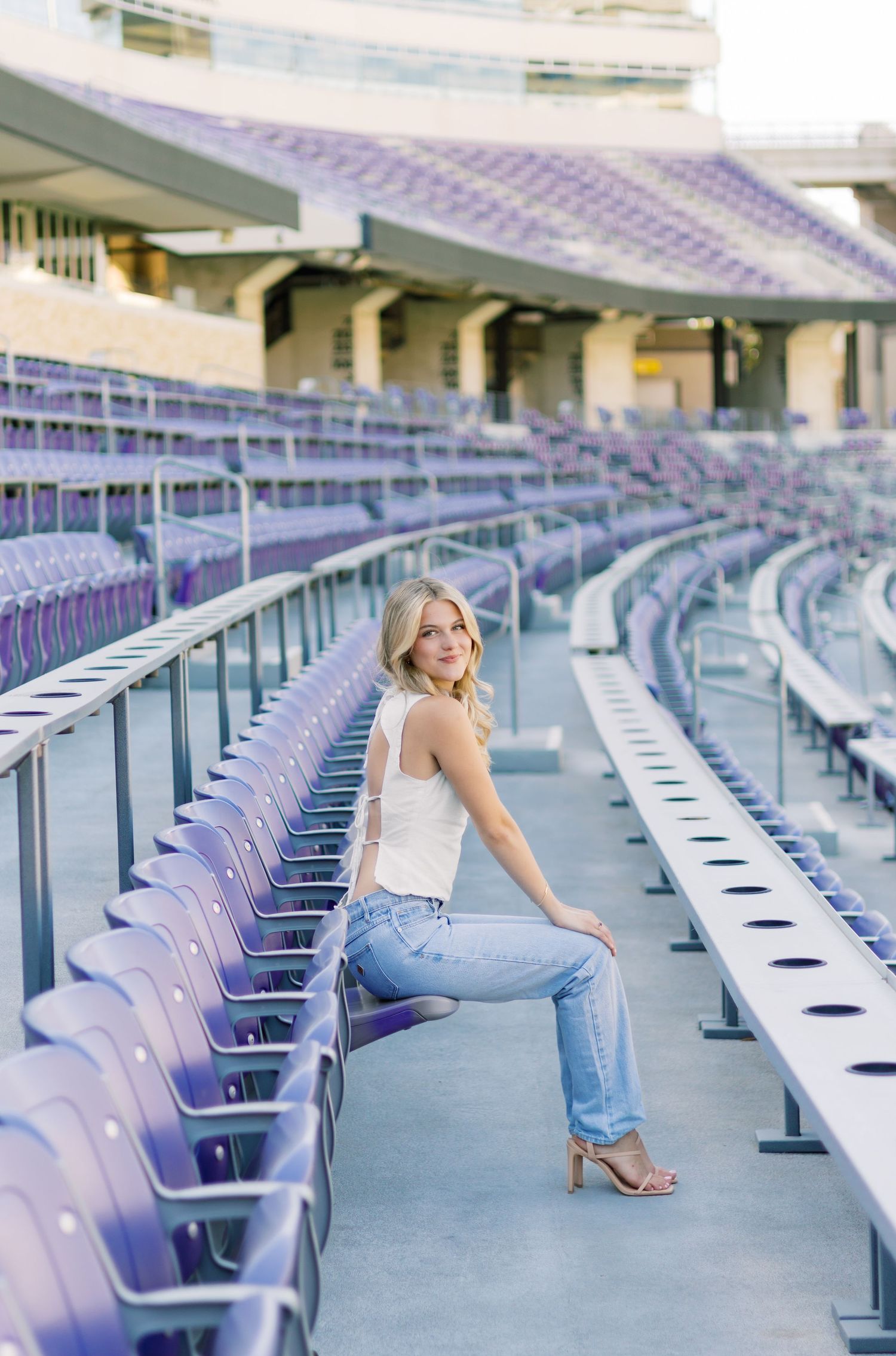Blonde woman in white tank and jeans posing on purple stadium bleachers with heels.