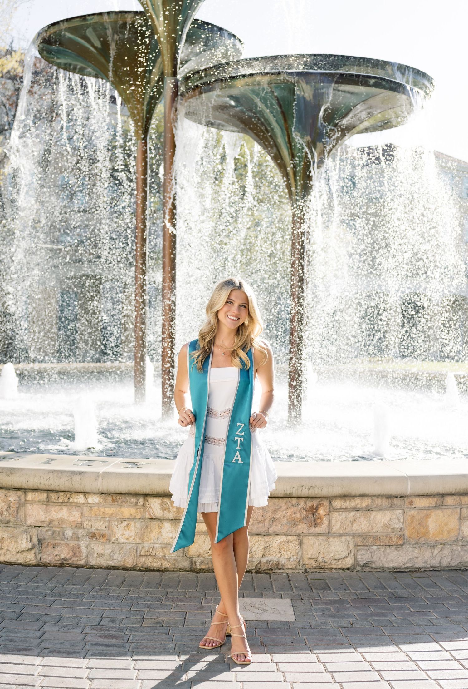 Smiling blonde woman in white dress wears teal ZTA graduation sash in front of decorative fountain.
