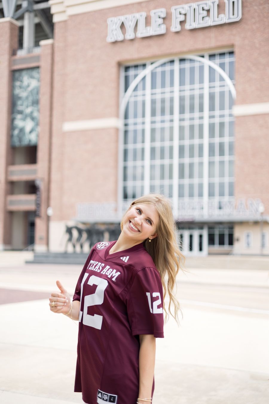 Smiling blonde woman in Texas A&M jersey giving thumbs up outside Kyle Field stadium.