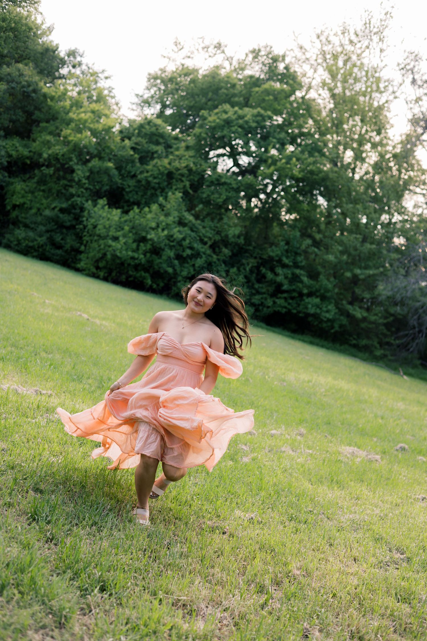 Young woman in peach off-shoulder dress running through lush green meadow, hair flowing in wind.