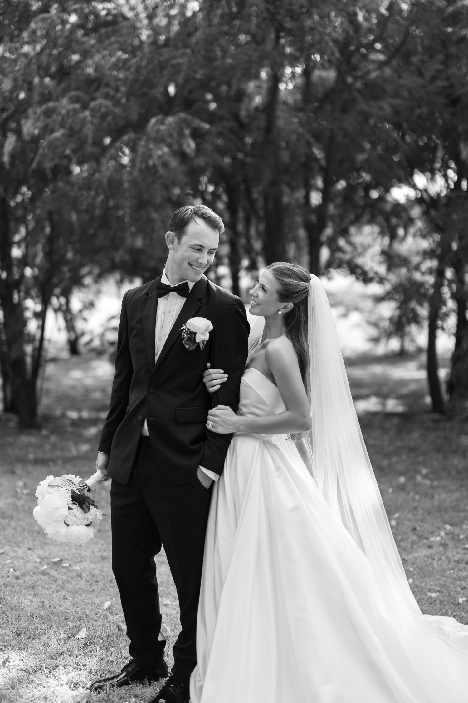 Black and white photo of smiling bride and groom embracing outdoors among trees on their wedding day.