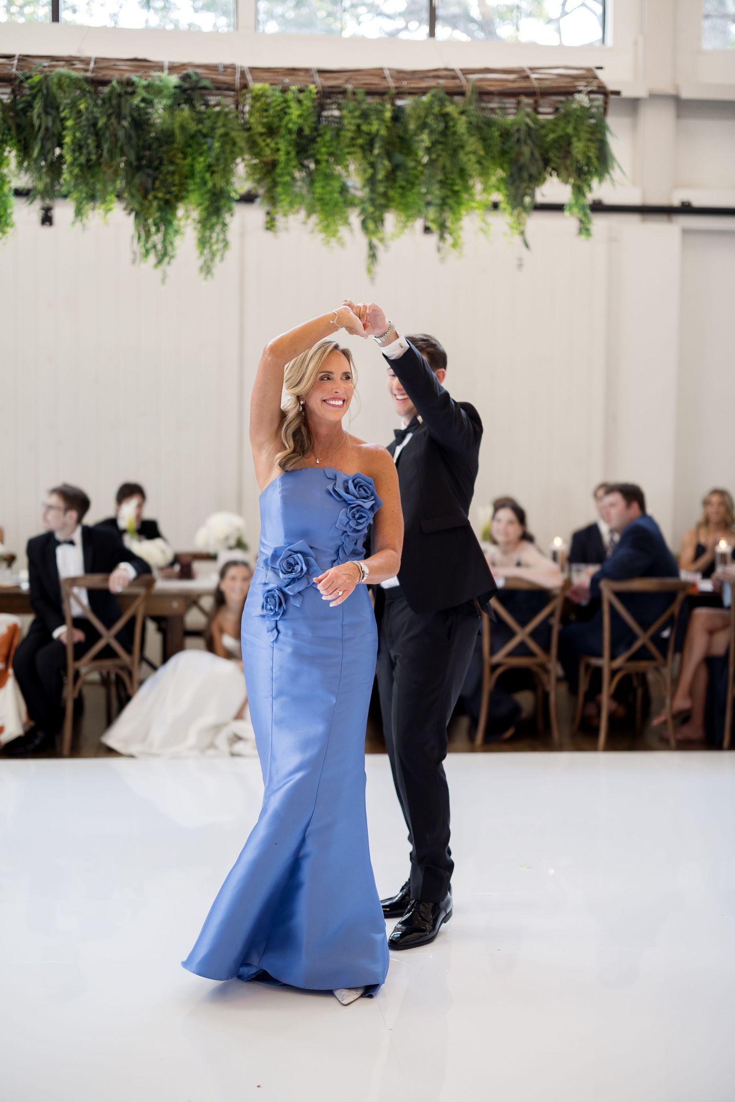 Mother and son share elegant first dance, she in blue floral gown, at a wedding reception with greenery overhead.