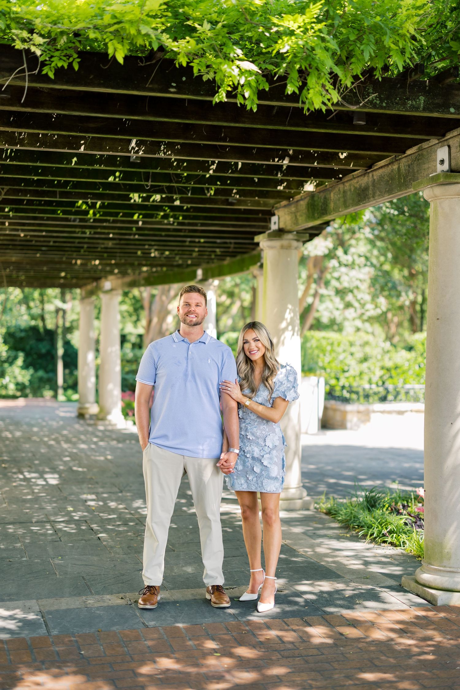 Couple posing under a vine-covered pergola in a lush garden, man in blue polo and woman in floral dress.