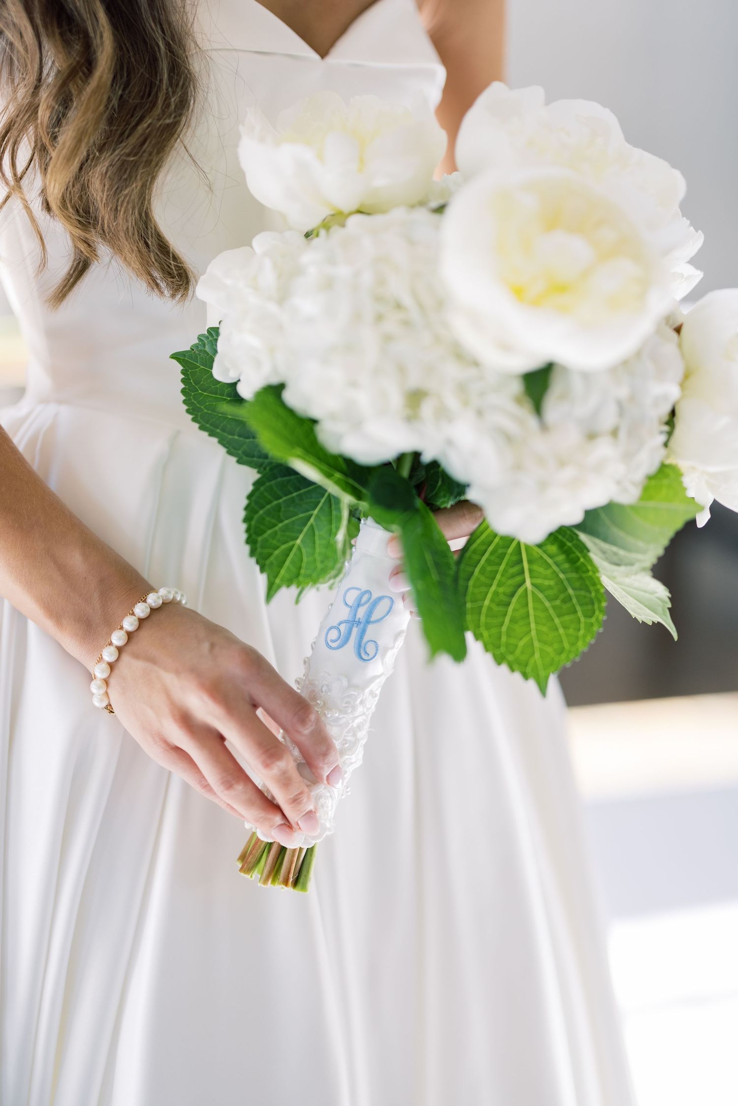 Bride holding white peony and hydrangea bouquet with monogrammed ribbon wrap and pearl bracelet.