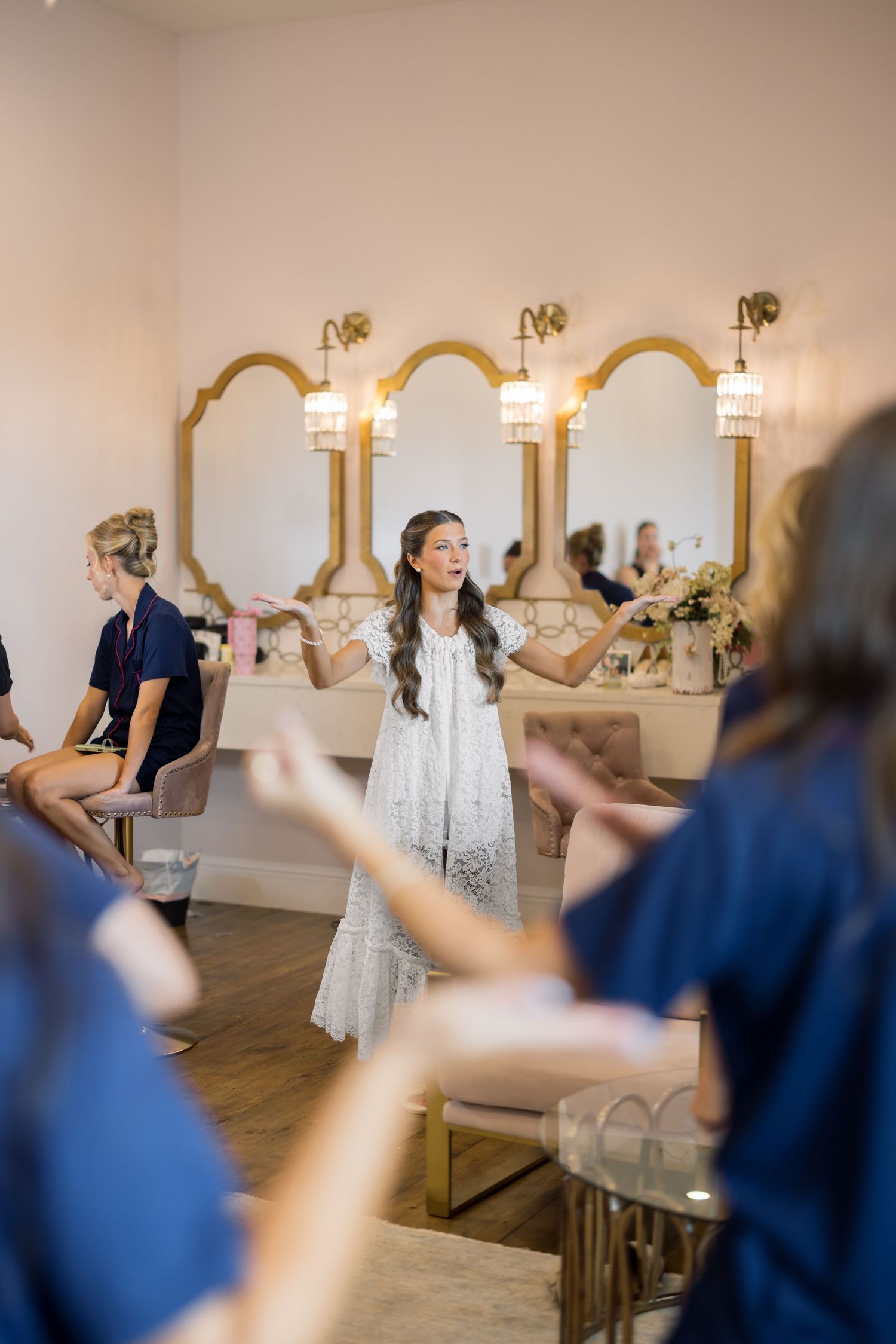 Bride in white lace dress stands in elegant bridal suite with gold mirrors, surrounded by bridesmaids in navy robes.