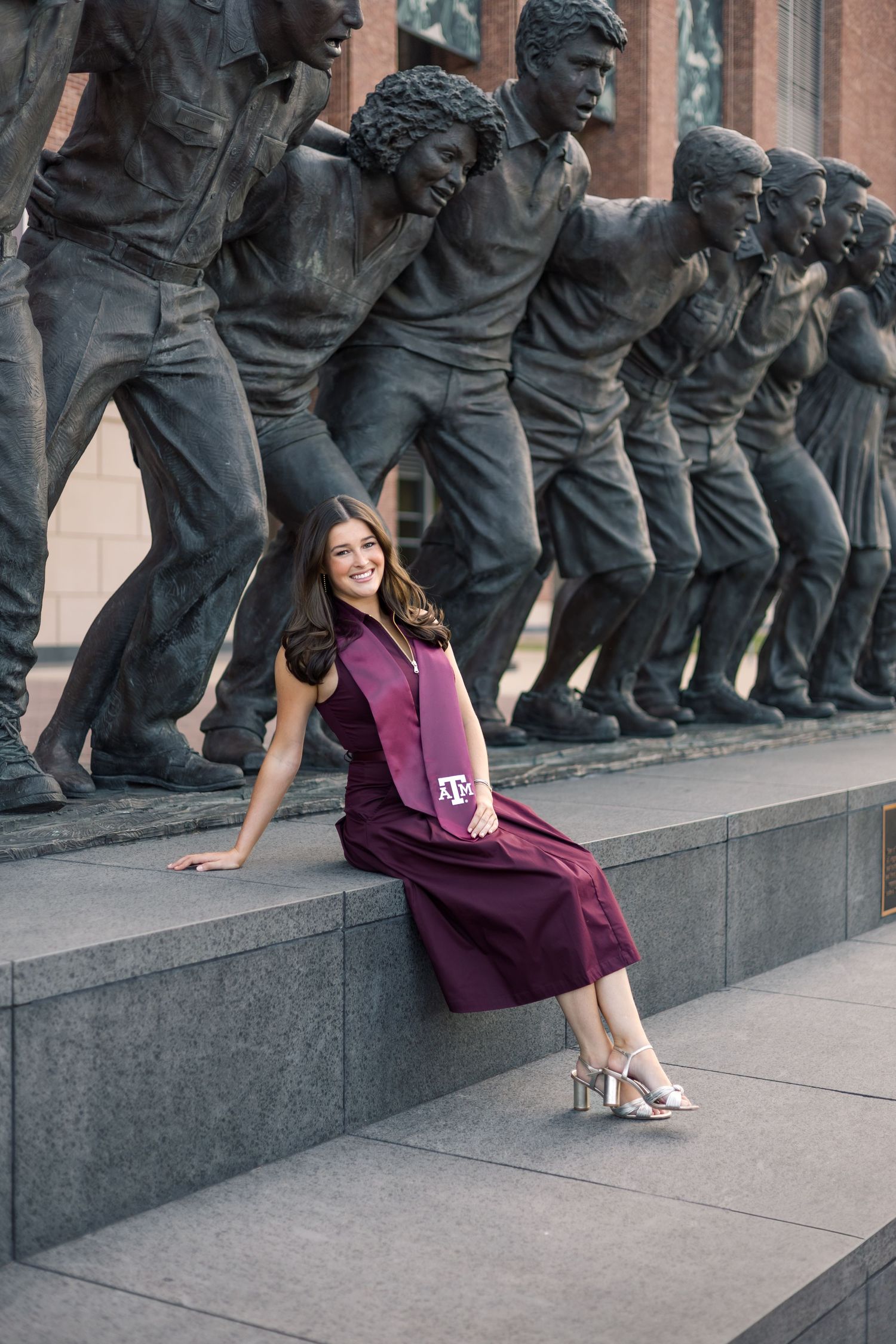 Texas A&M graduate in maroon dress poses on steps beside bronze statues at Kyle Field.