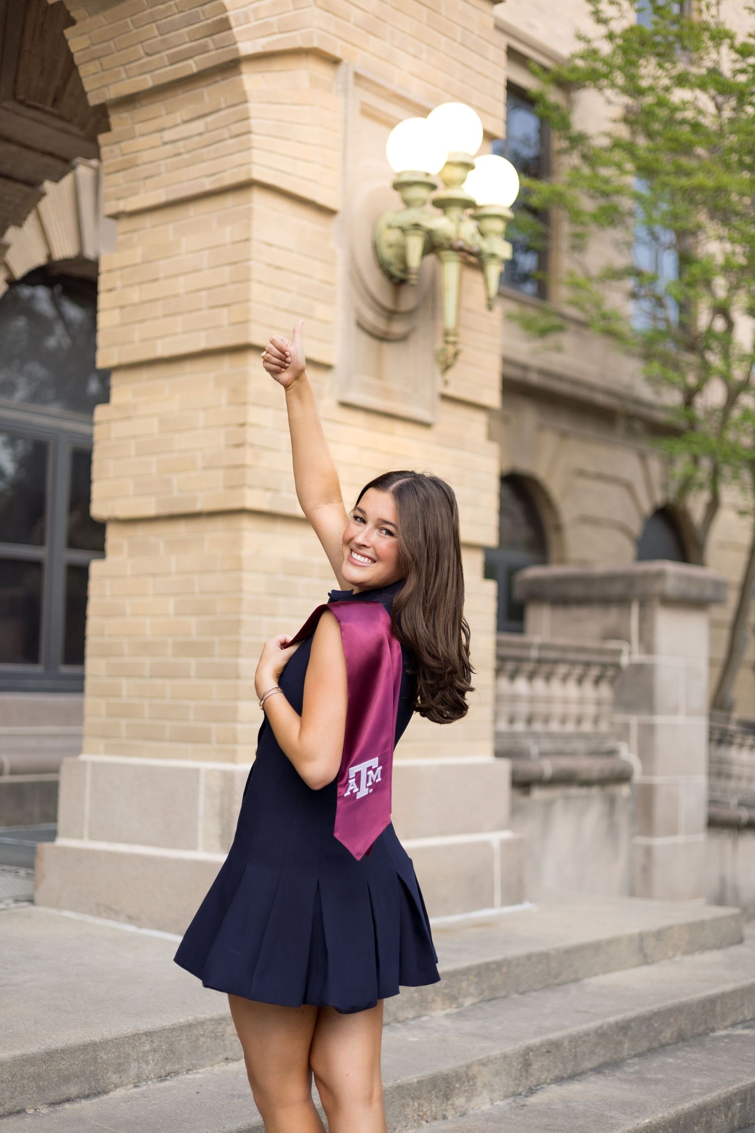 Joyful Texas A&M graduate in navy dress raising arm in celebration outside a stone building.