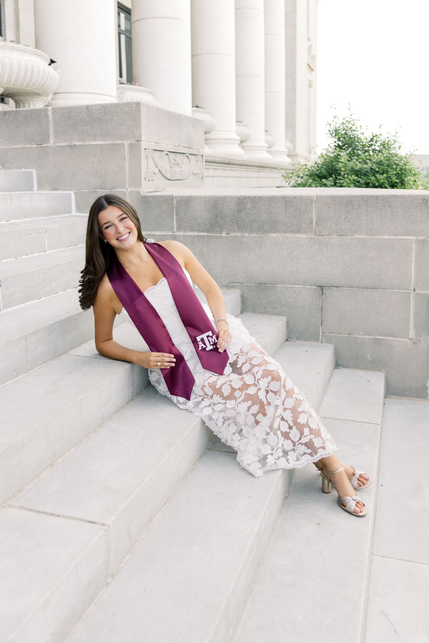 Joyful woman in white floral lace dress with A&M graduation stole posing on marble stairs.