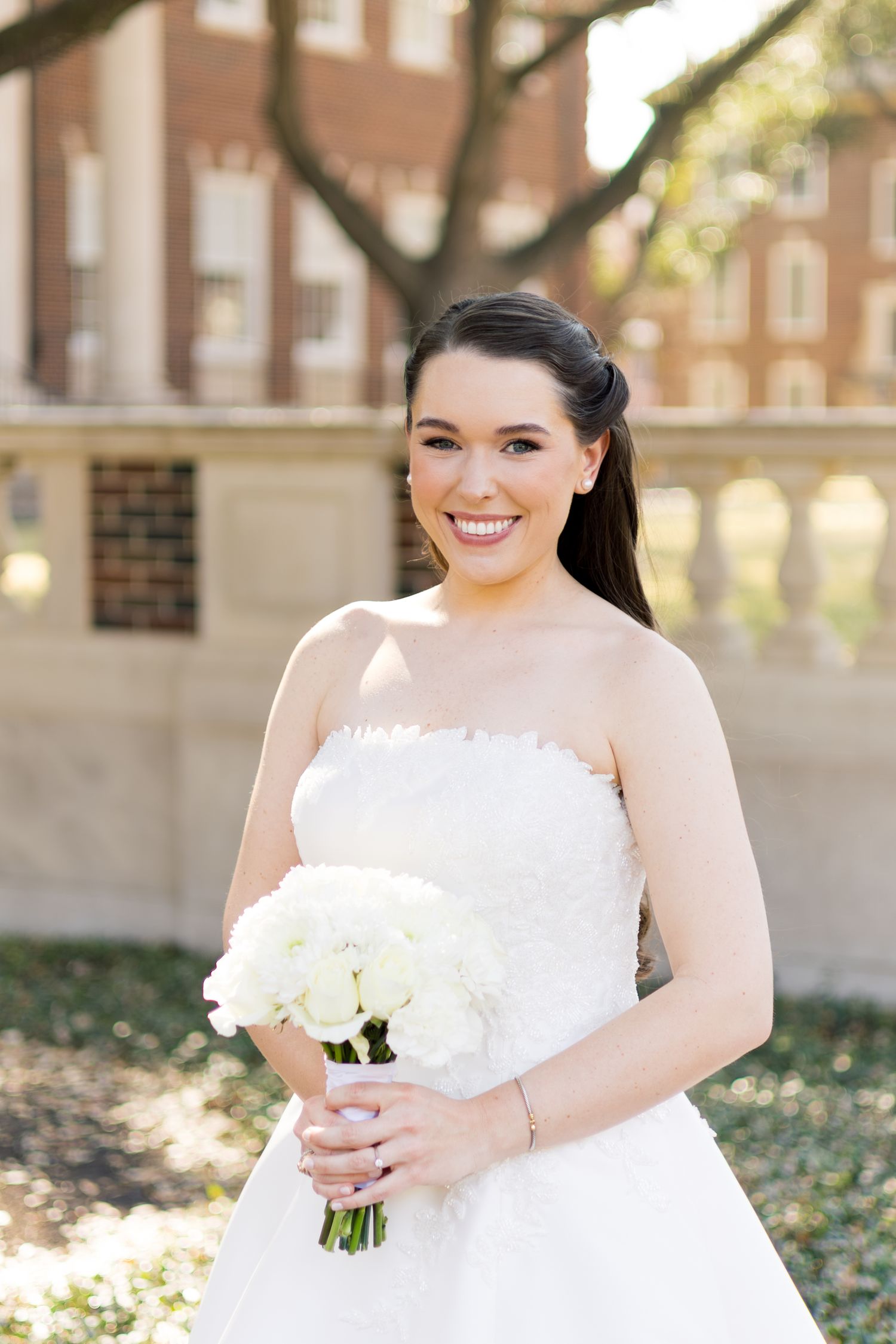 Radiant bride in a strapless white gown holds a bouquet of white roses outdoors.