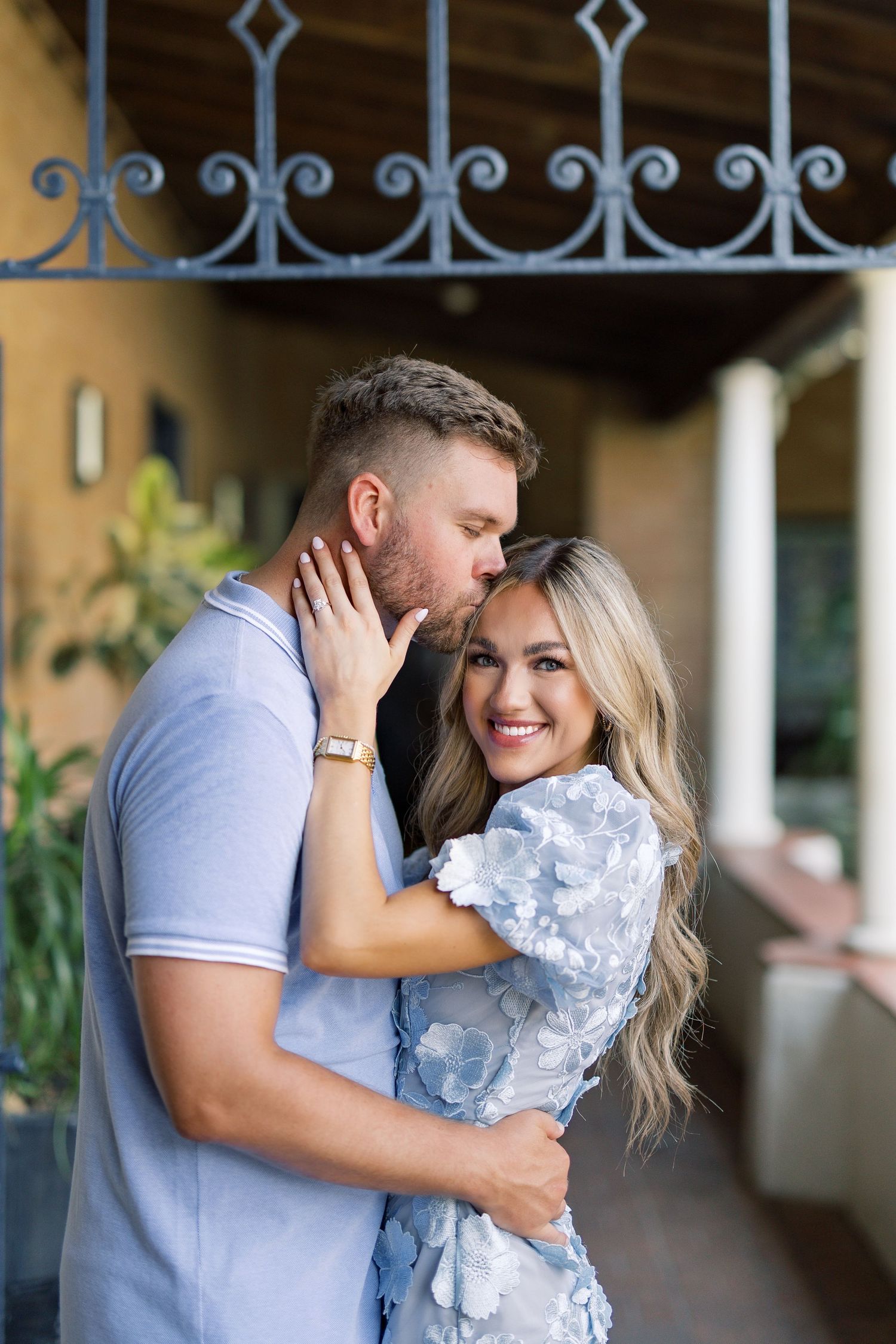 A couple embraces and smiles on a porch, the man kissing her temple, both dressed in light blue outfits.