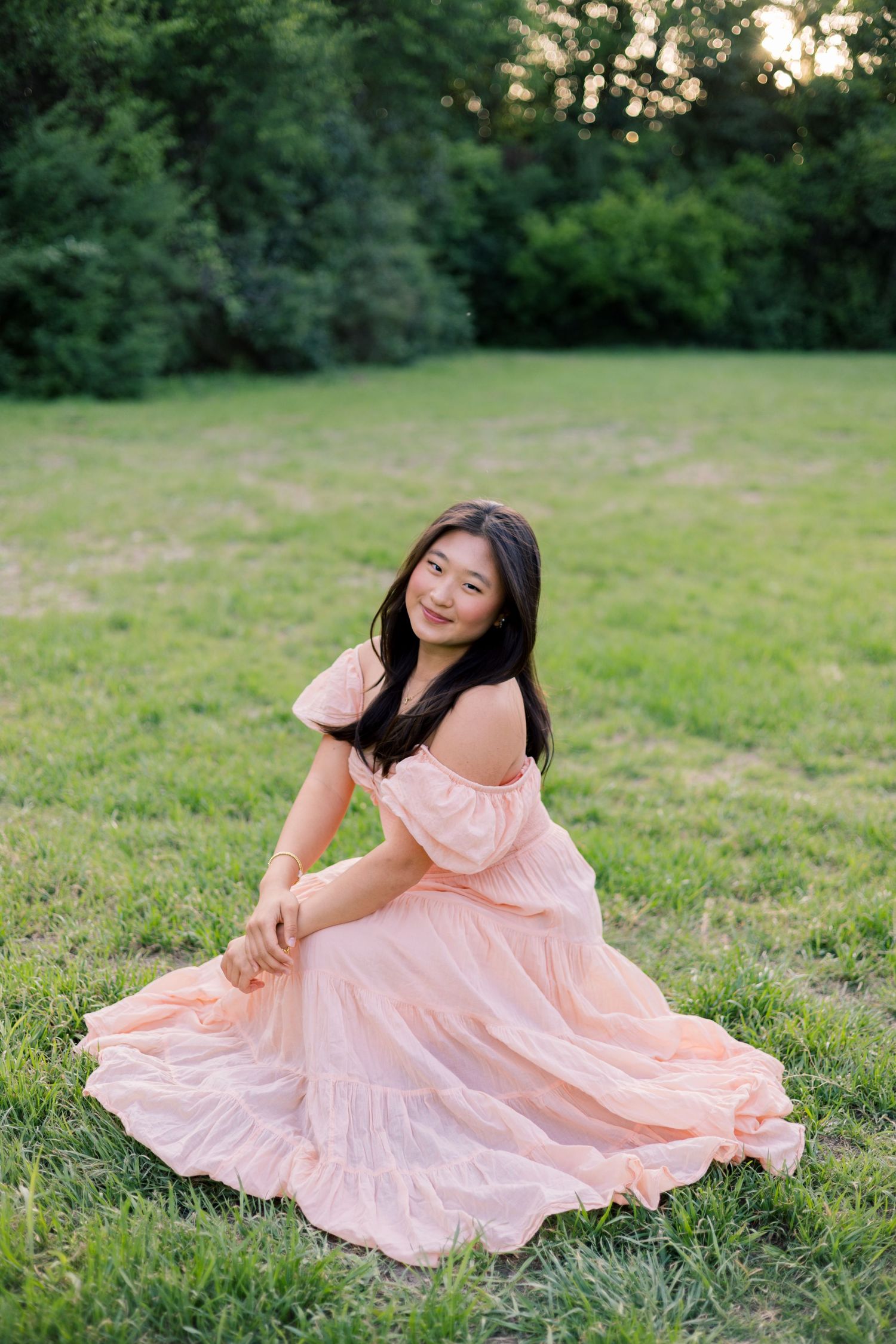 Young Asian woman in a pink off-shoulder dress sitting gracefully on green grass with trees in the background.