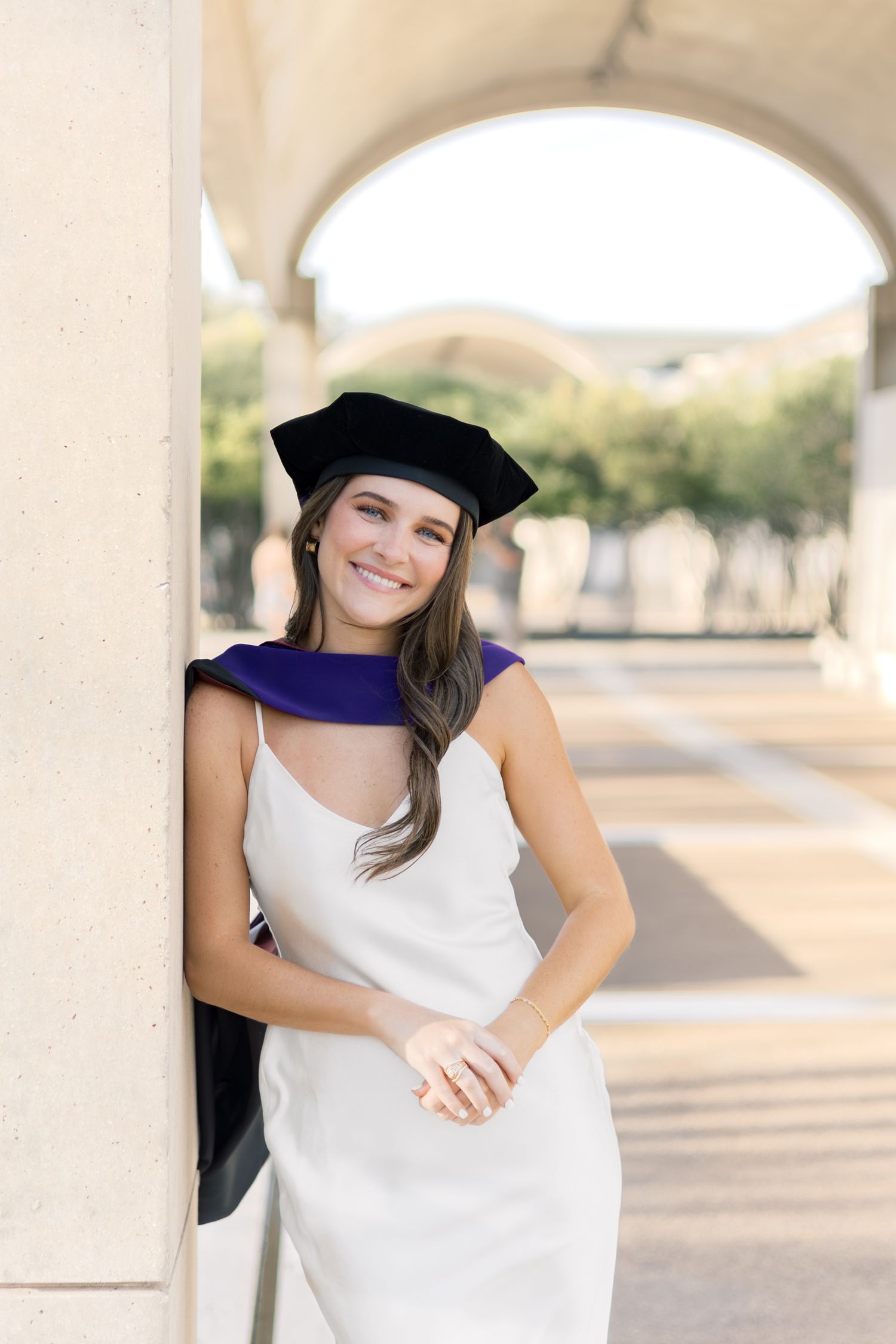 Smiling graduate woman in white dress and black doctoral cap with purple hood leaning against a column.