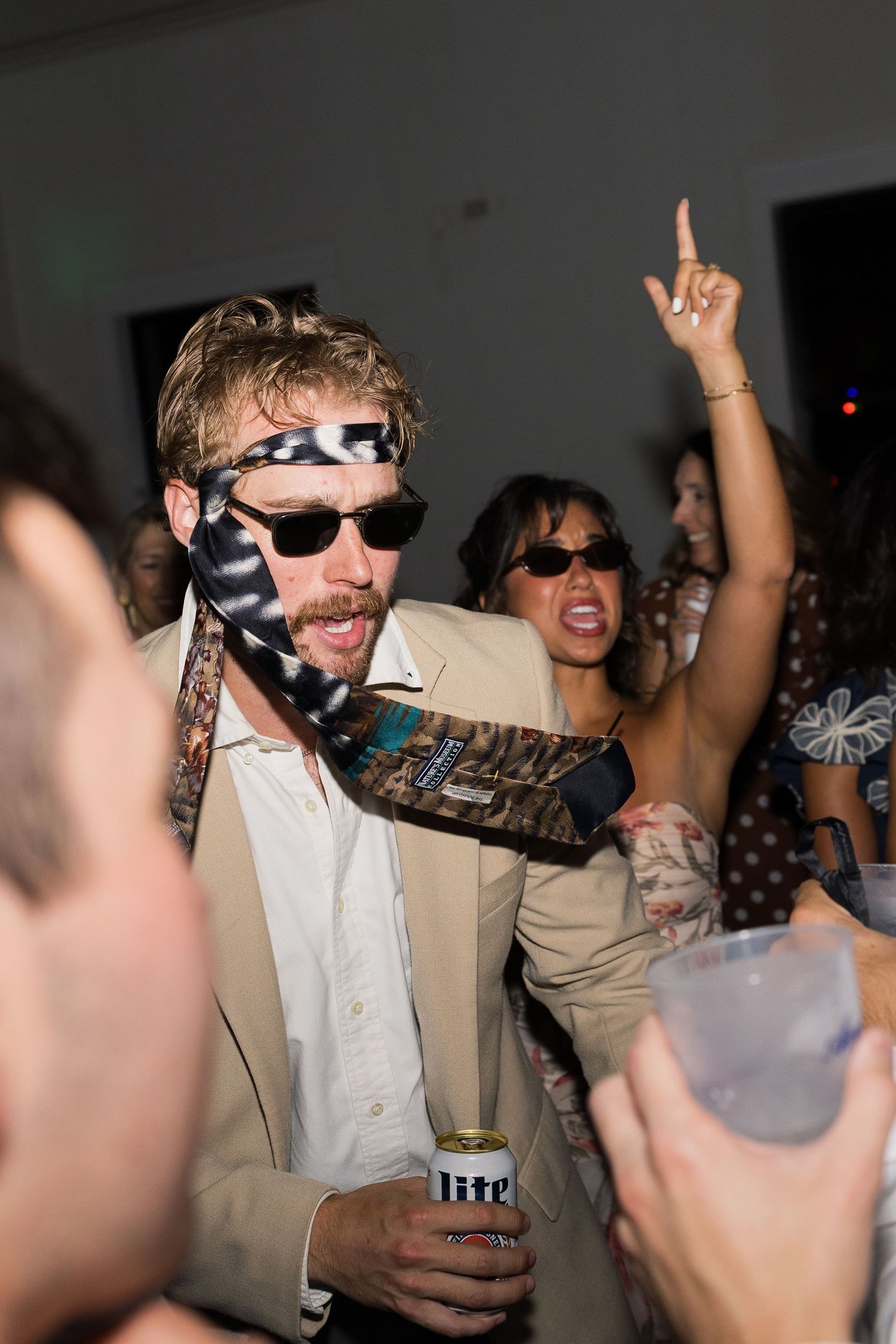 Man with tie around head holding Miller Lite beer at lively party with dancing guests in background.