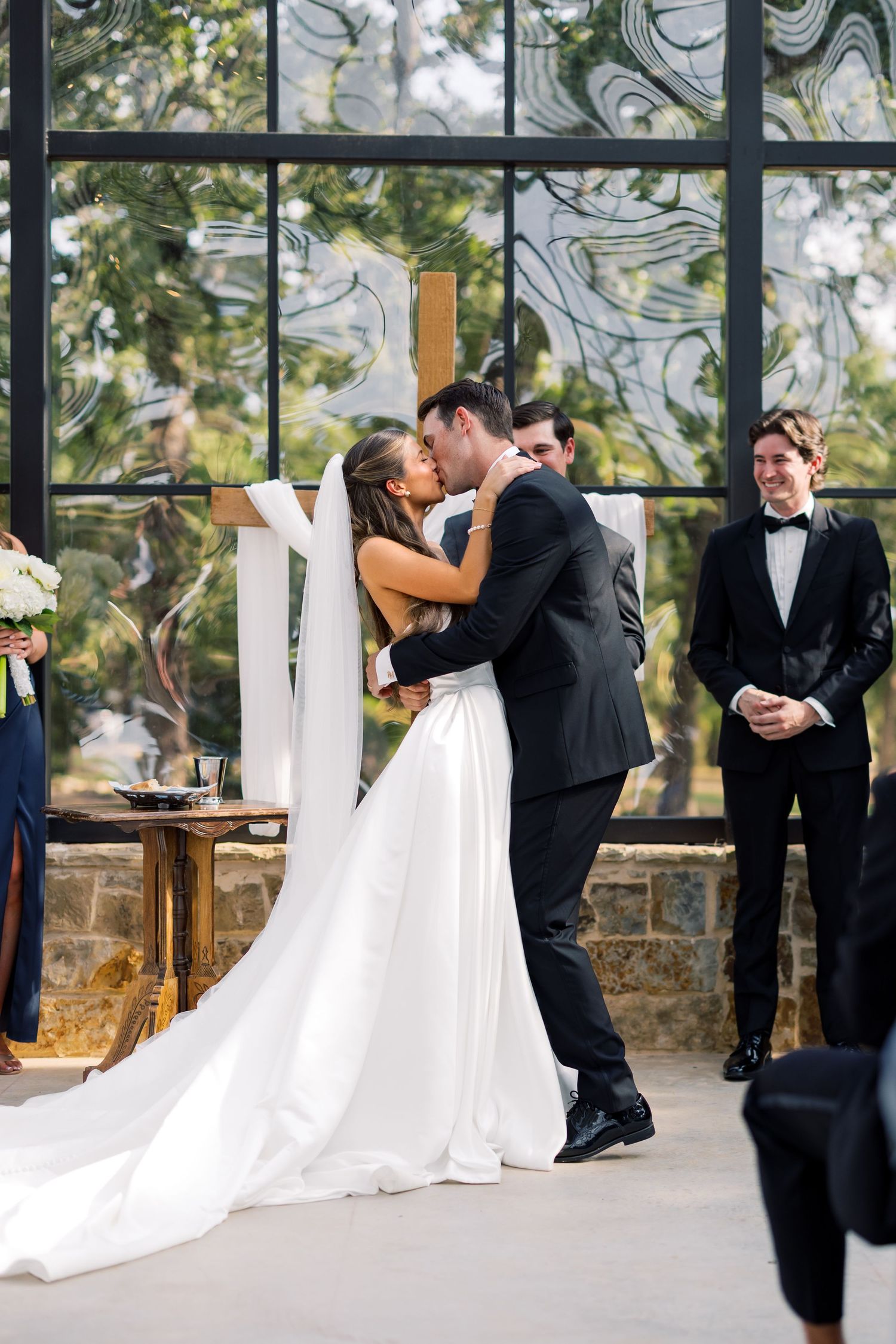Bride and groom share first kiss at outdoor wedding ceremony with elegant glass windows and wood cross backdrop.