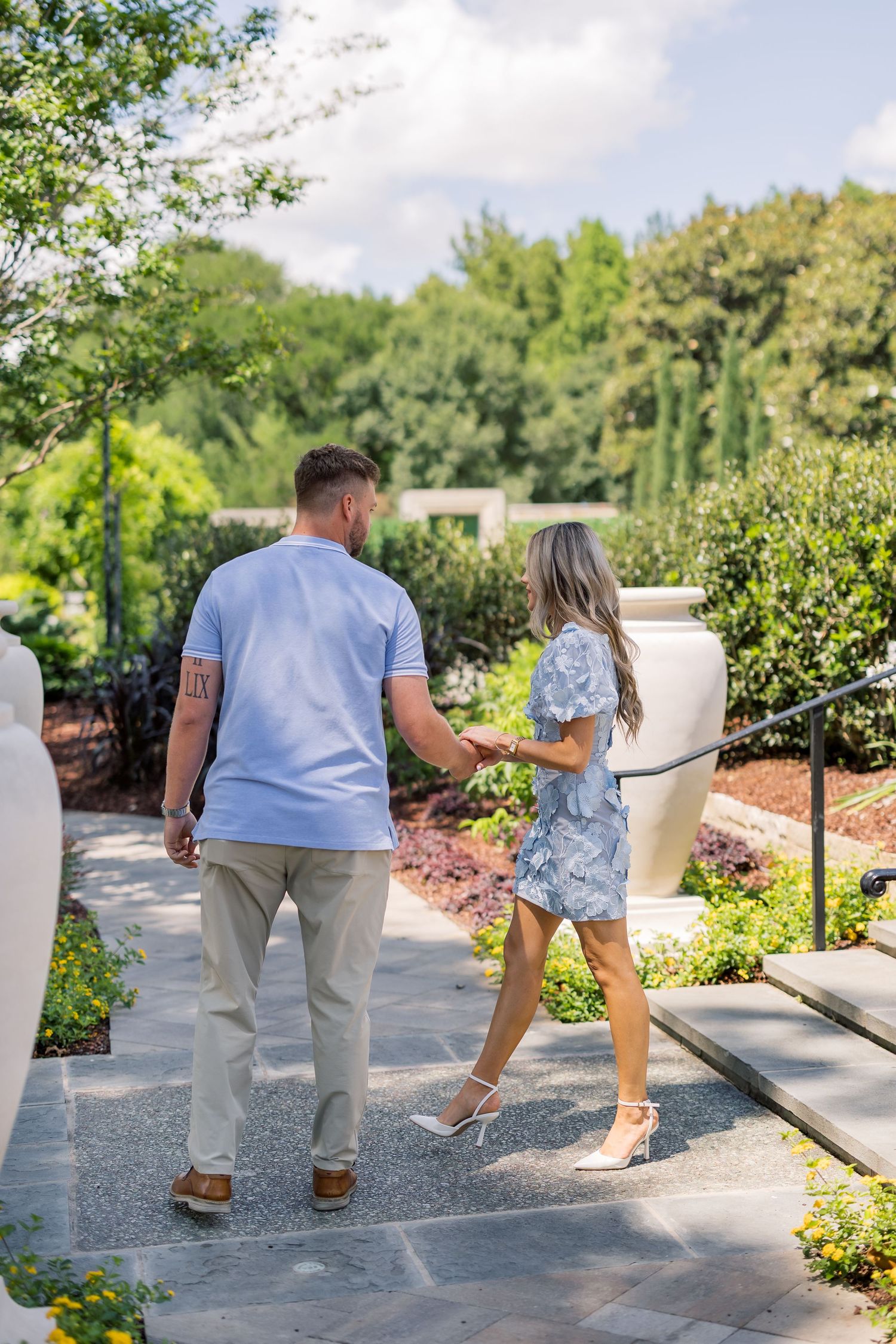A couple holds hands walking through a lush garden, woman in floral dress and heels, man in blue polo and khakis.