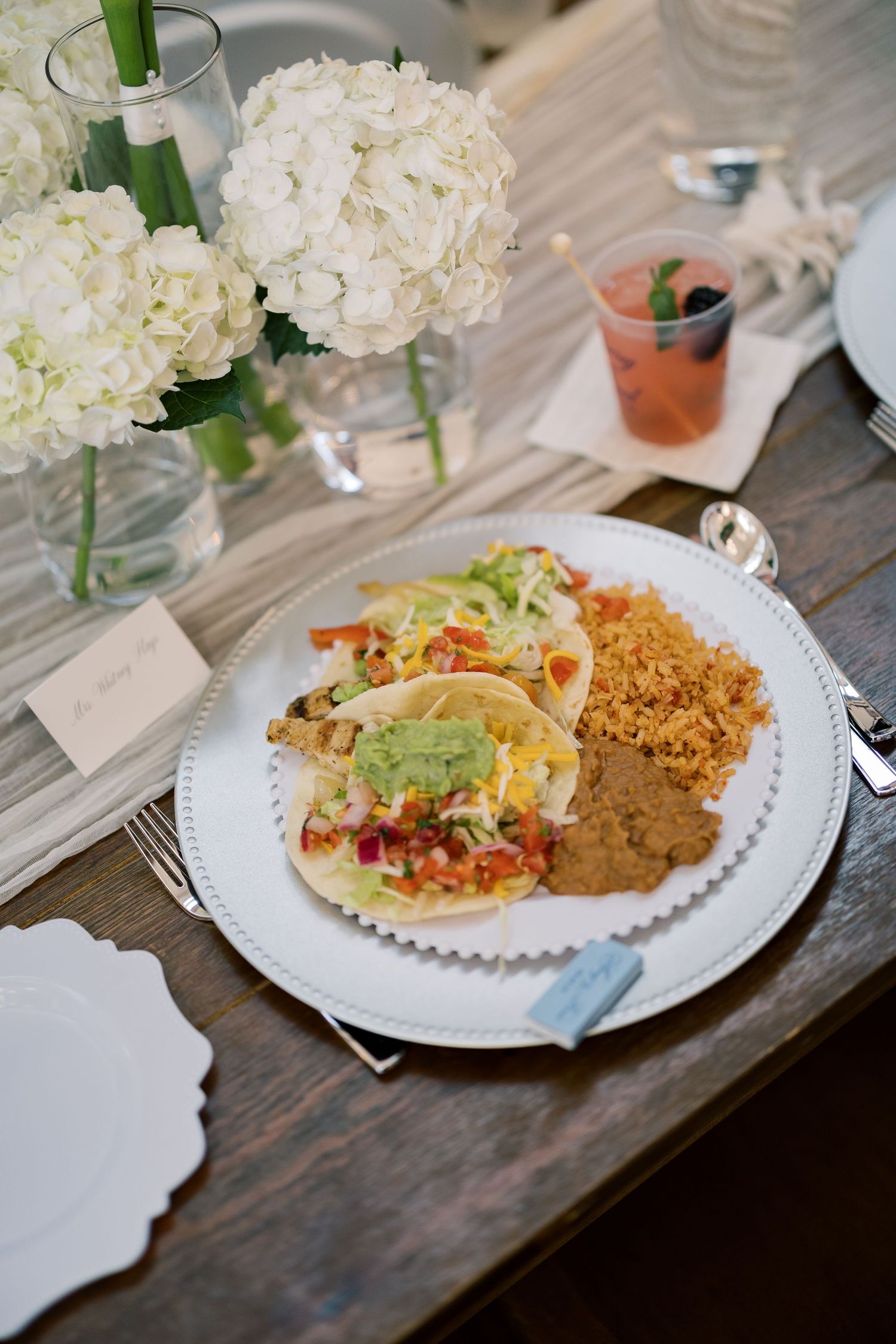 Elegant wedding reception plate with fish tacos, rice, and beans beside white hydrangea centerpieces on a rustic wood table.