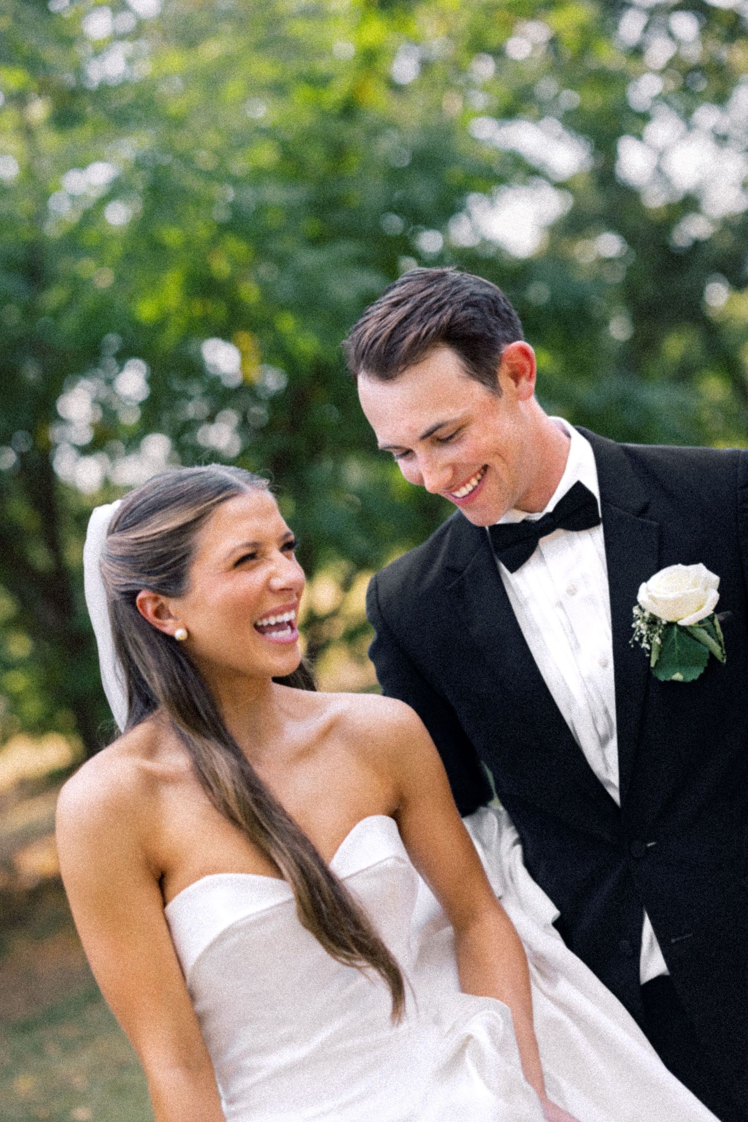 Smiling bride in strapless white gown and groom in black tuxedo with white boutonniere share a joyful moment outdoors.