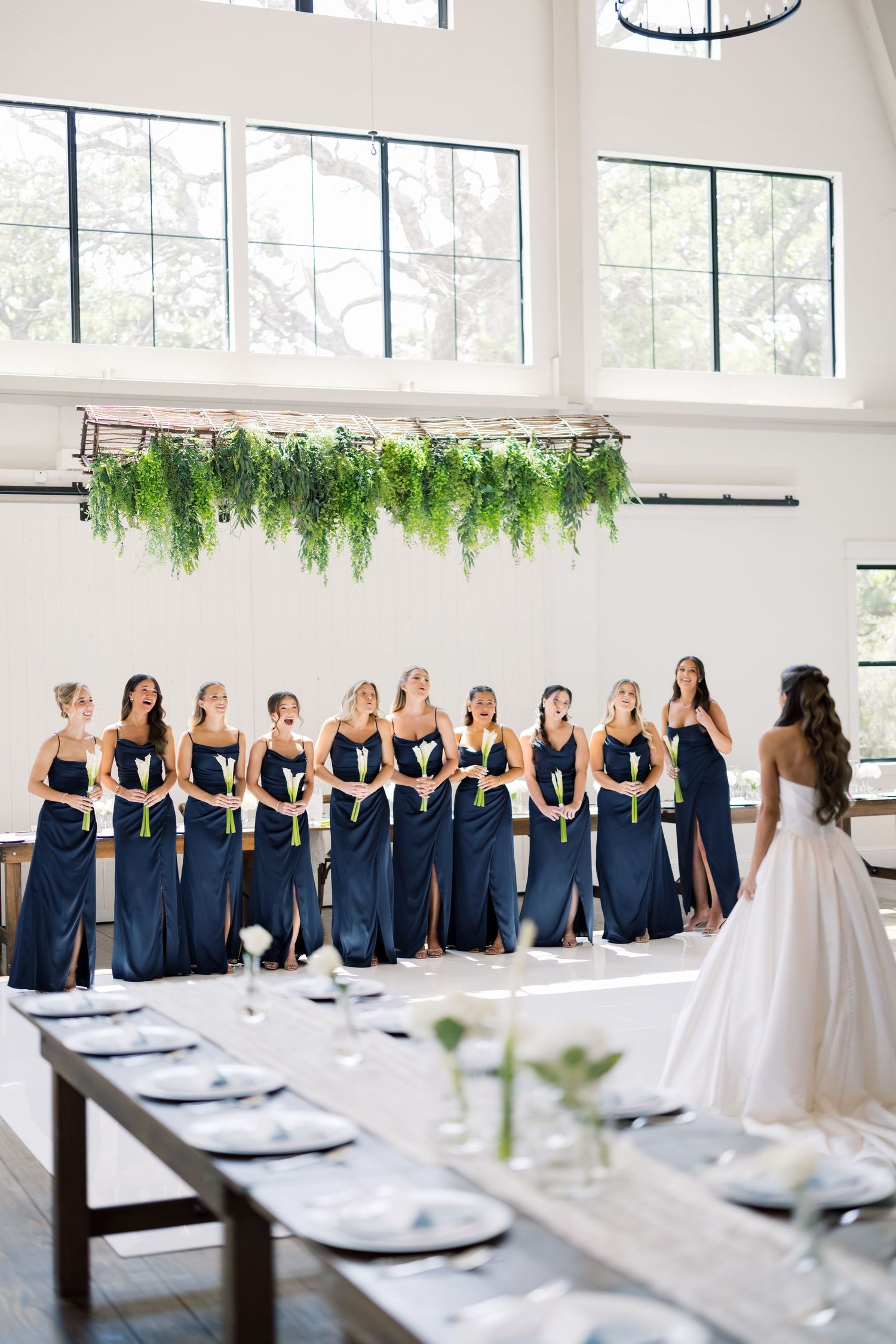 Bride faces navy blue bridesmaids holding white flowers in bright white venue with hanging greenery installation.