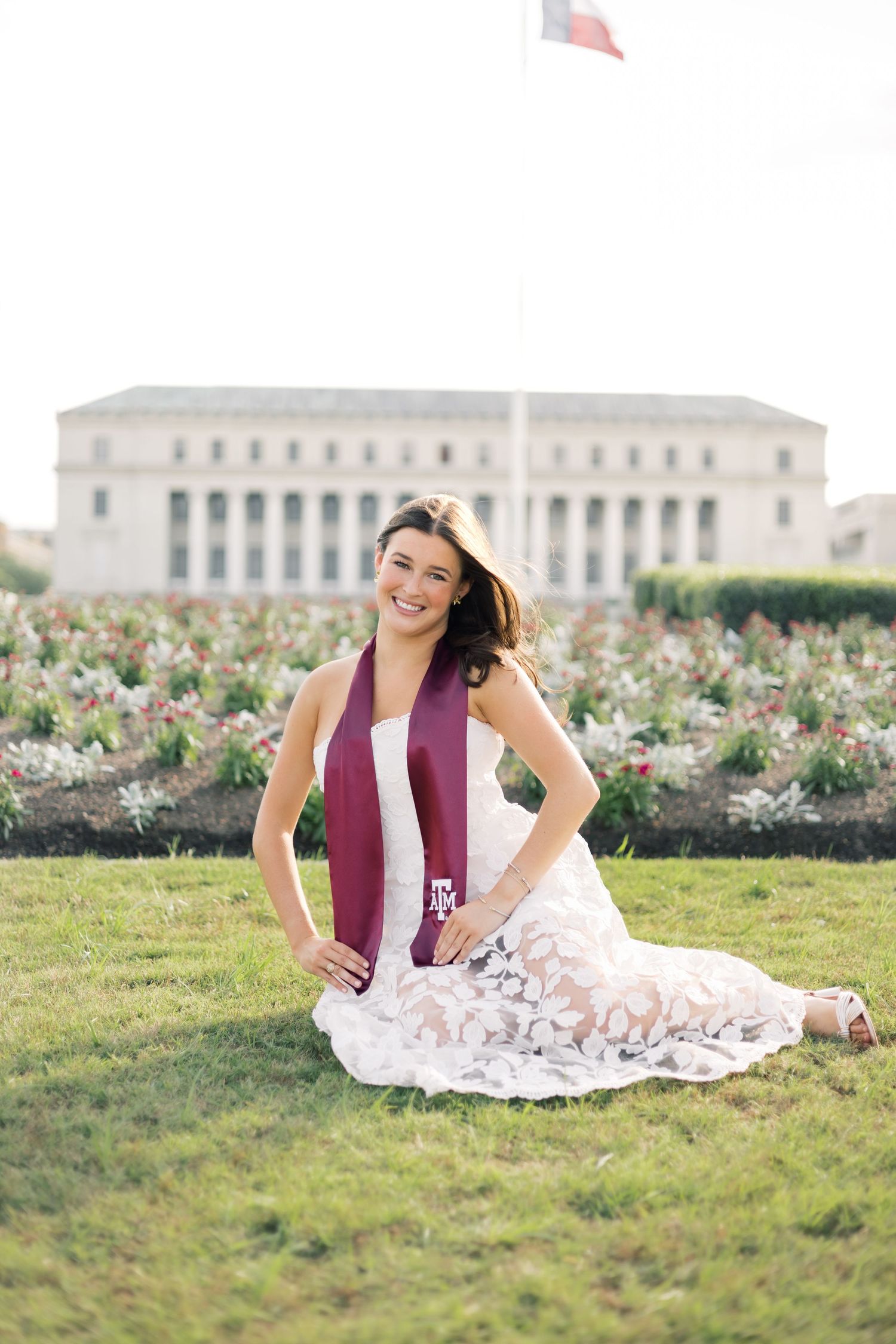 Smiling woman in white lace dress with maroon Texas A&M graduation stole sitting on grass at Texas A&M University.