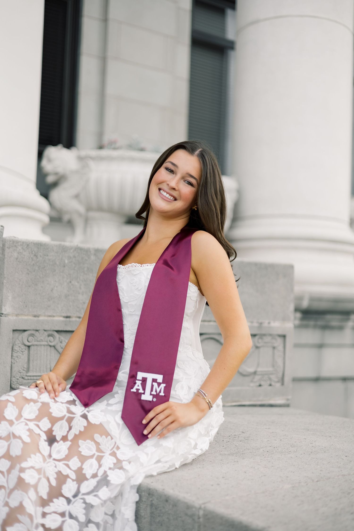 Smiling woman in white floral lace dress with Texas A&M maroon graduation stole seated on stone steps by columns.
