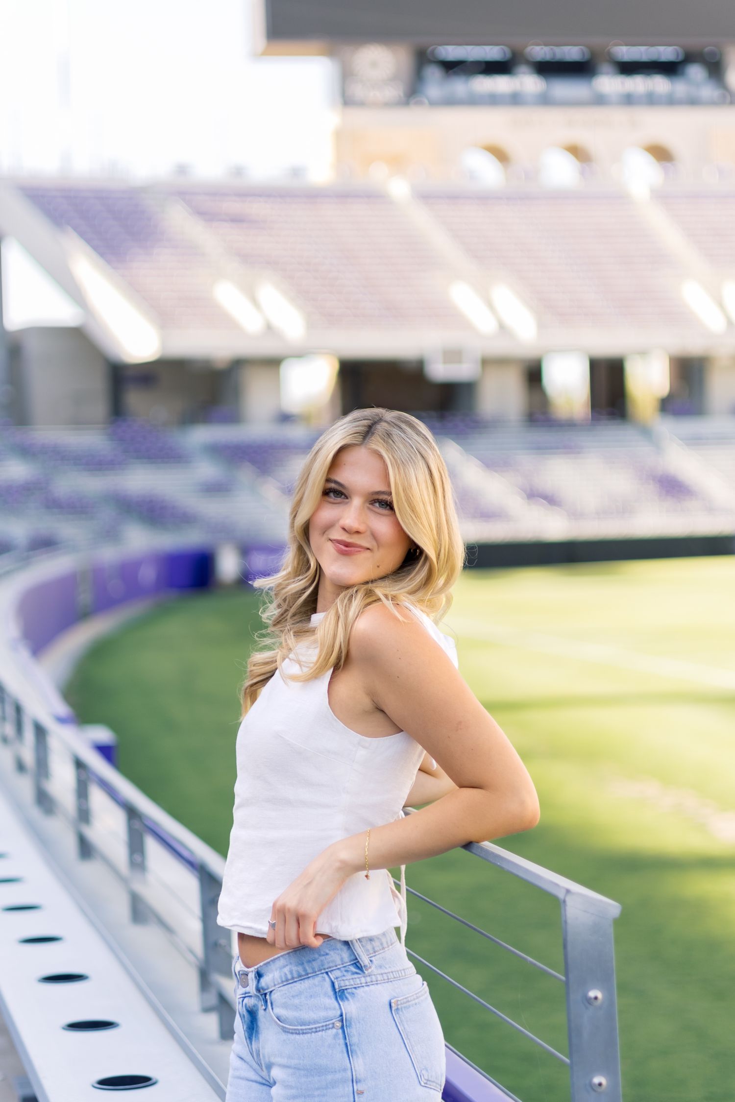 Smiling blonde woman in white tank top and jeans leaning on railing at purple stadium.
